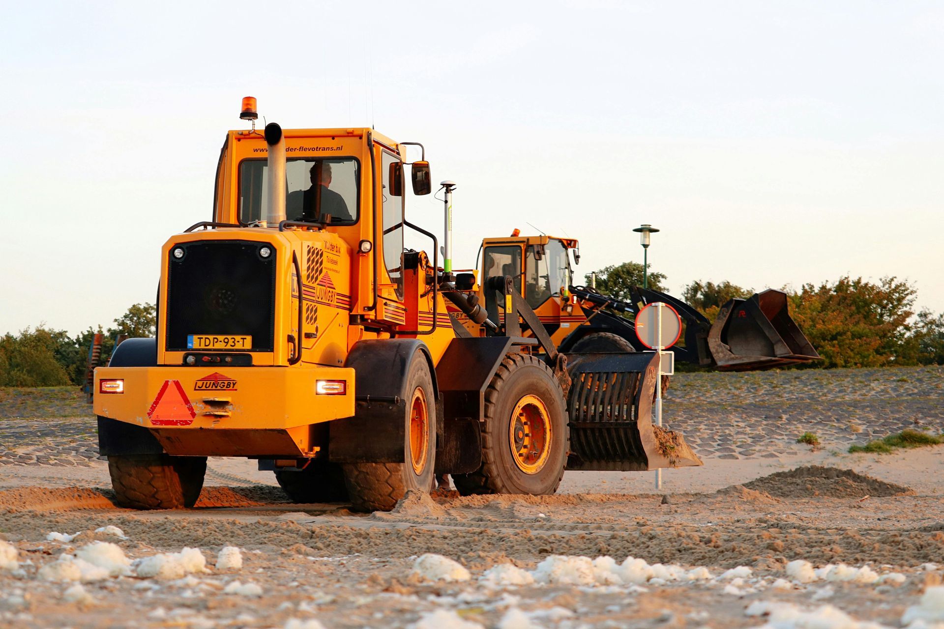 Yellow loader on gravel, operator visible, another loader in the background; outdoor construction site.