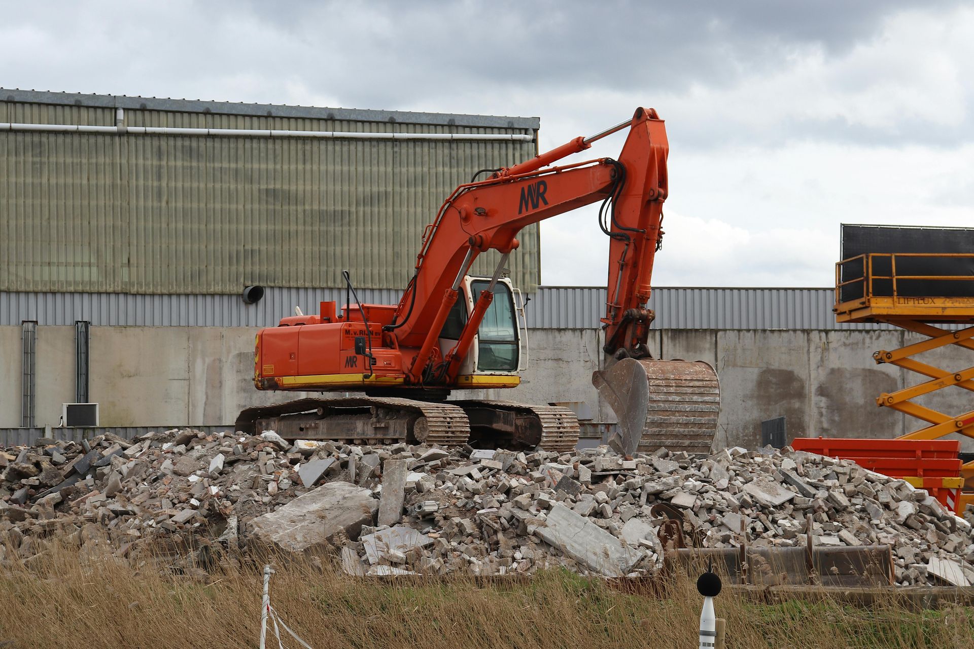 Orange excavator demolishing debris in front of a building on a cloudy day.