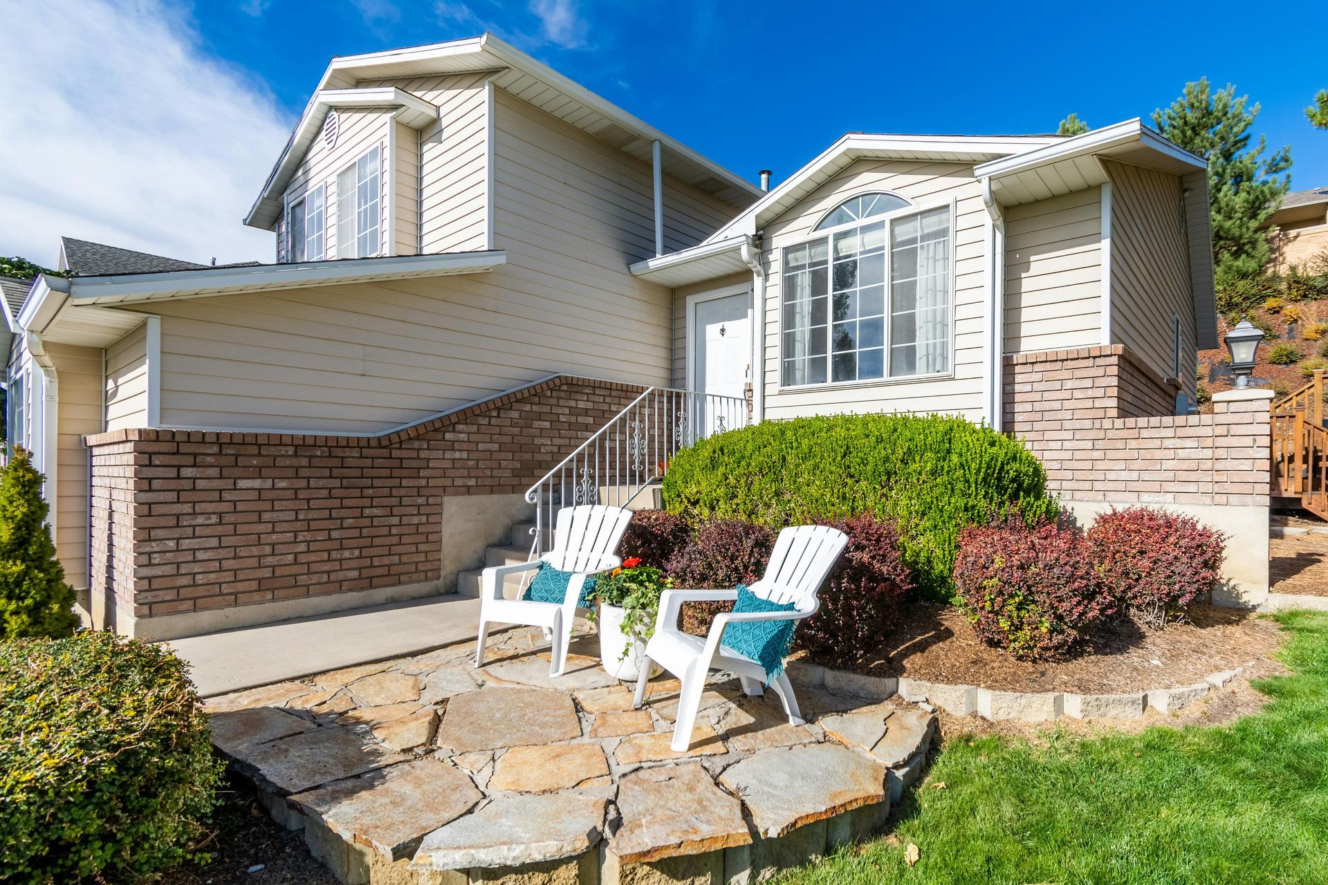 Two-story beige house with brick accents and two white chairs on a stone patio.