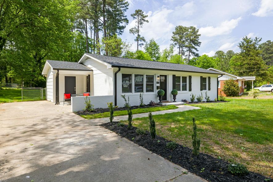 White ranch-style house with black trim and a concrete driveway. Green grass and trees surround the property.