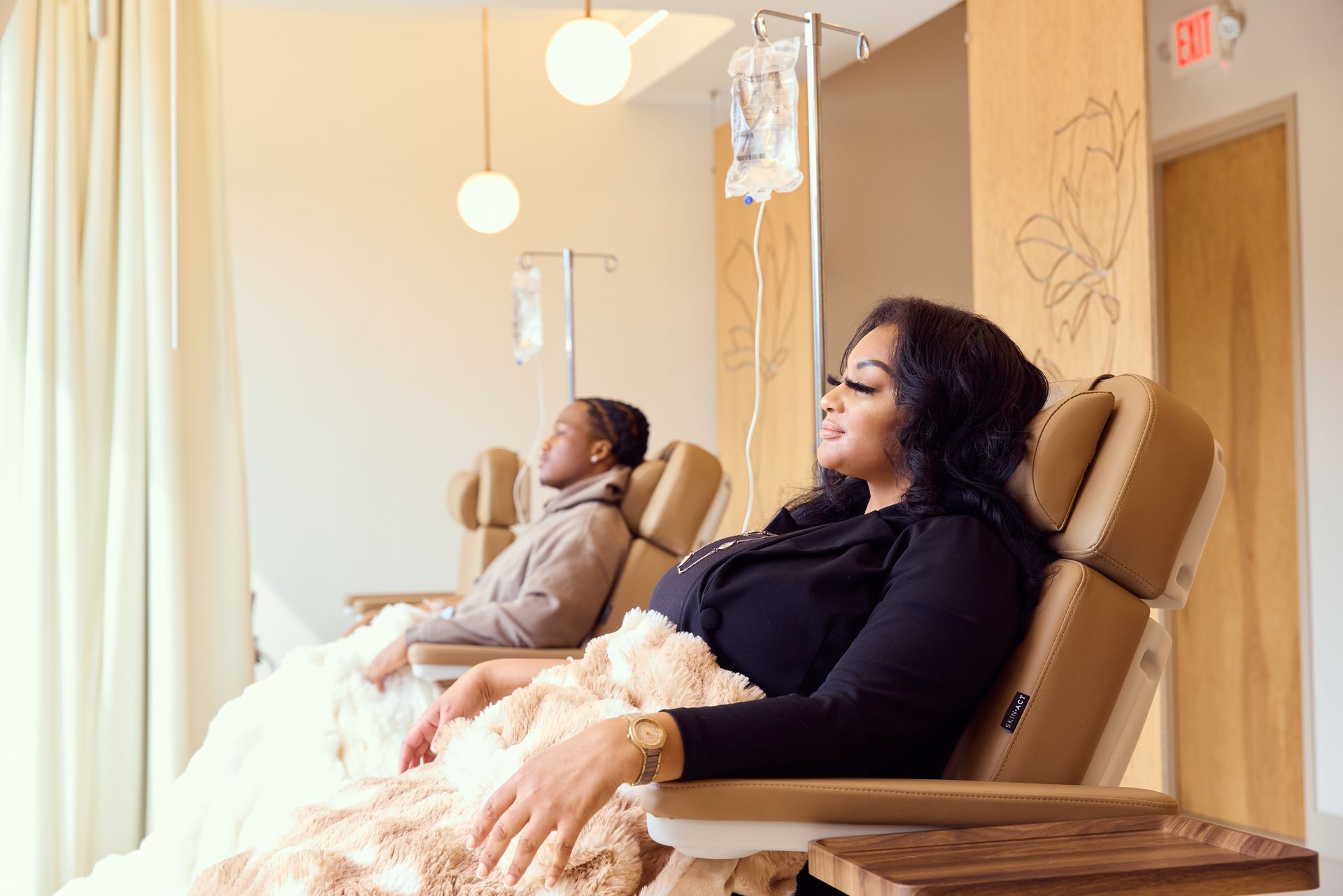 Two people receiving IV drips while seated in recliners in a bright room at Bain Beauty.