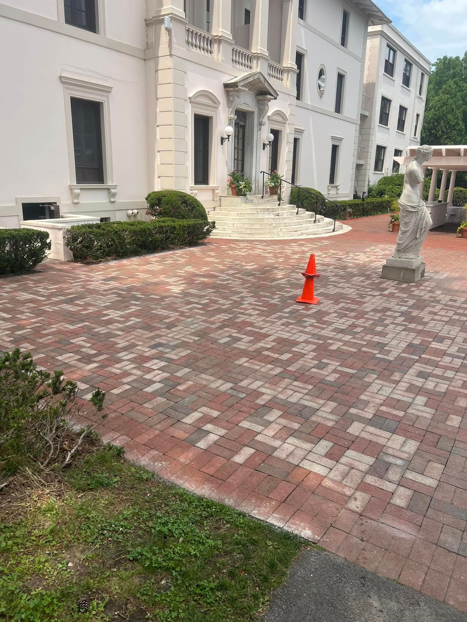 Brick paved driveway leading to a white building with a statue on the right. An orange cone is in the center.