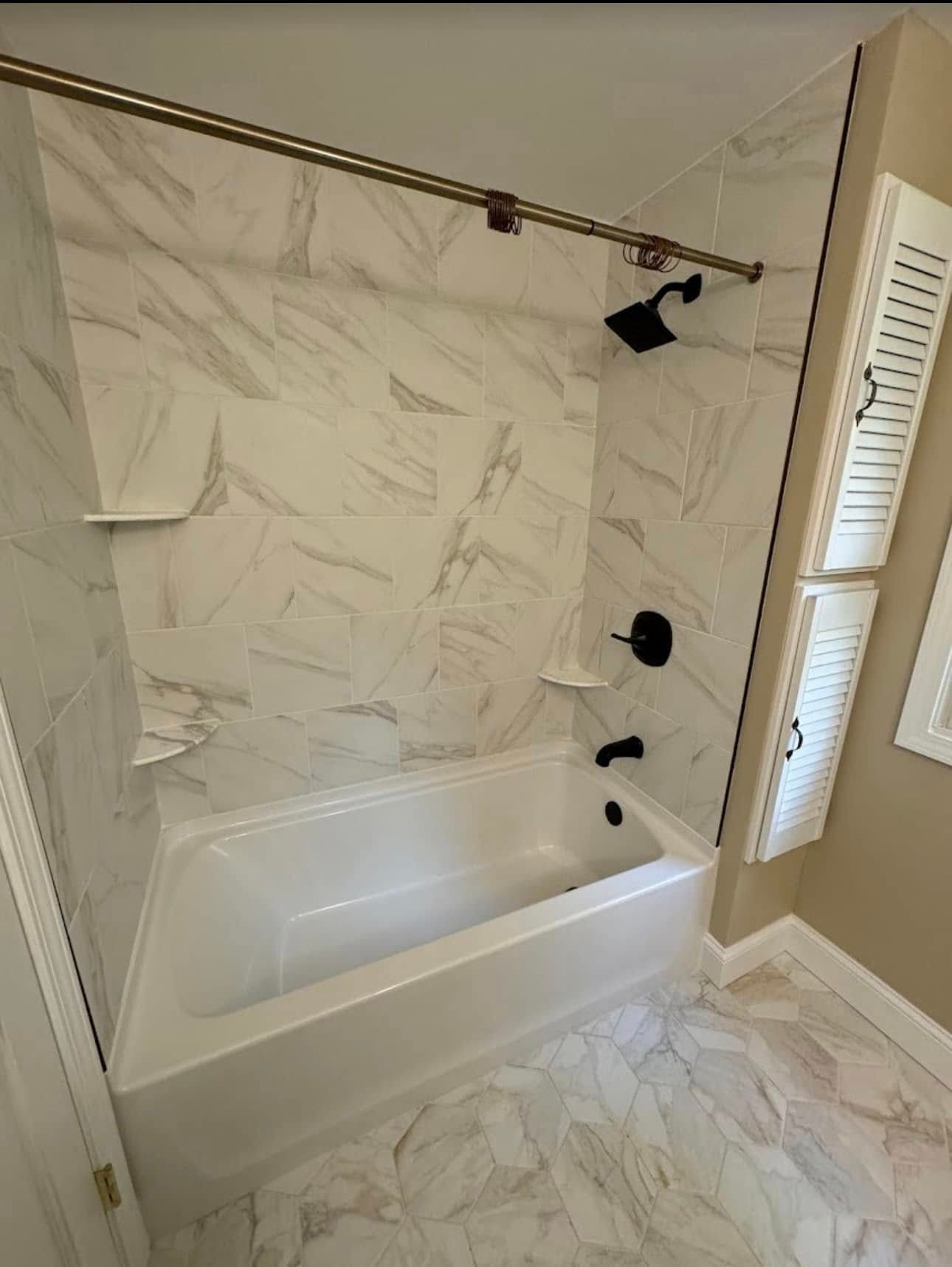 Bathroom with white tub, marble-look tile, and black fixtures; two white shutter-style cabinets on the wall.