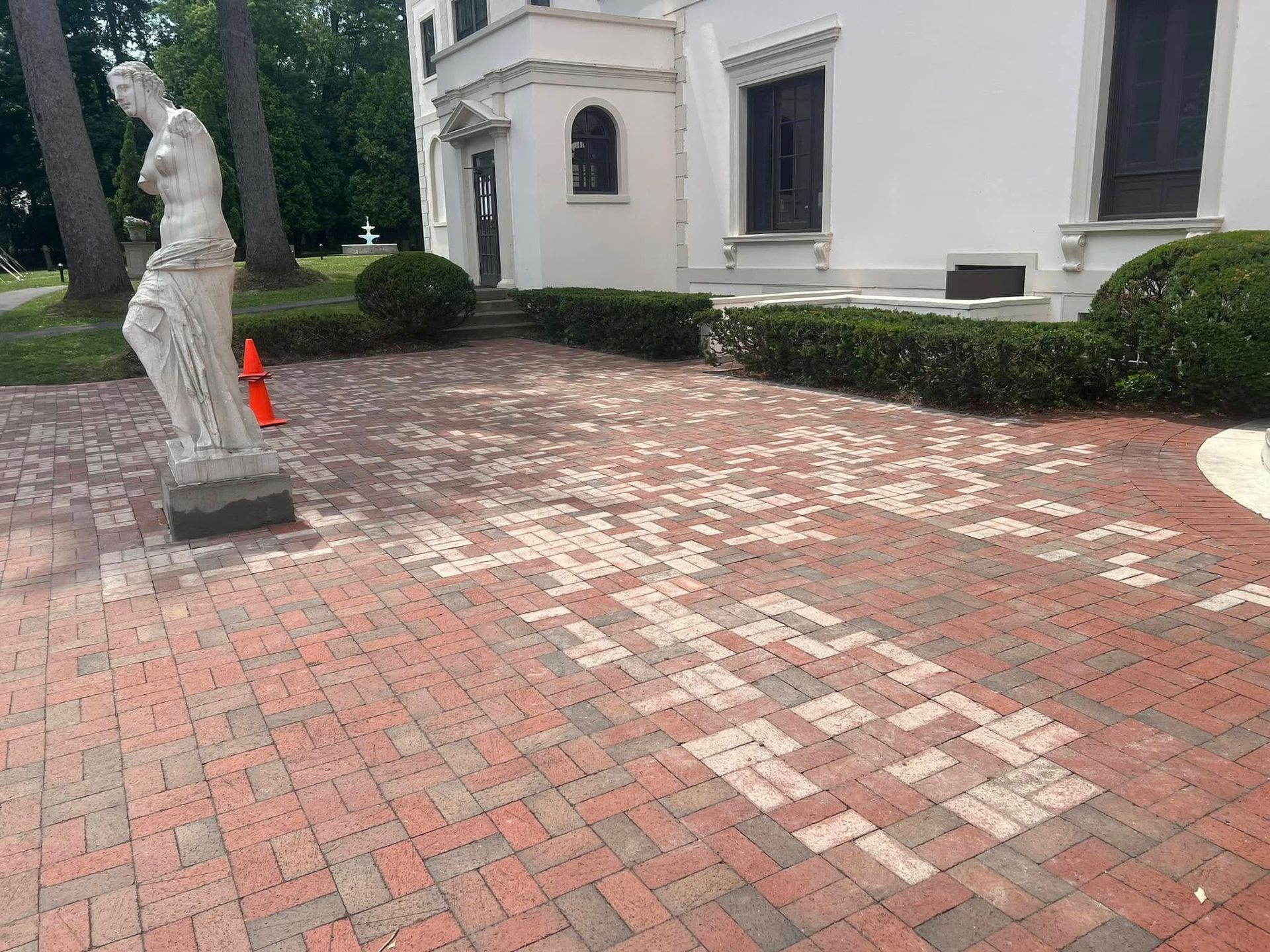Red brick patio with patchy discolored areas, statue, white building, and neatly trimmed hedges.