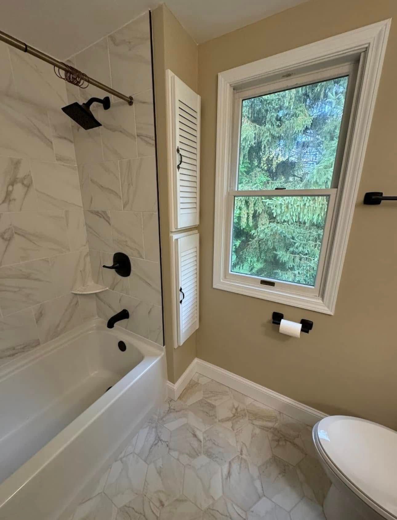 Bathroom with white and gray tiled walls, tub, window, and storage cabinet; neutral colors.