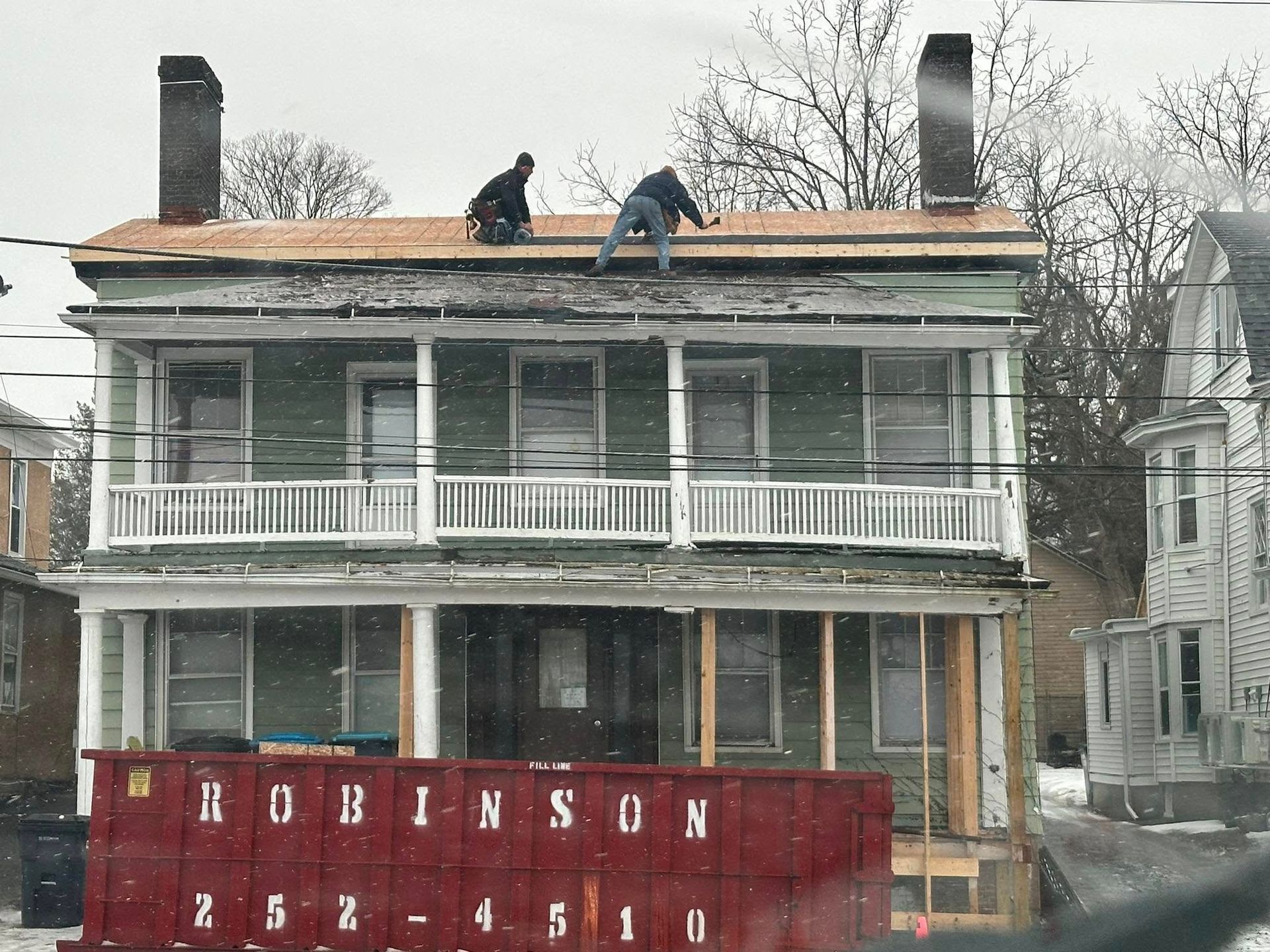 Two workers replacing a roof on a two-story green house in winter, red dumpster in front.