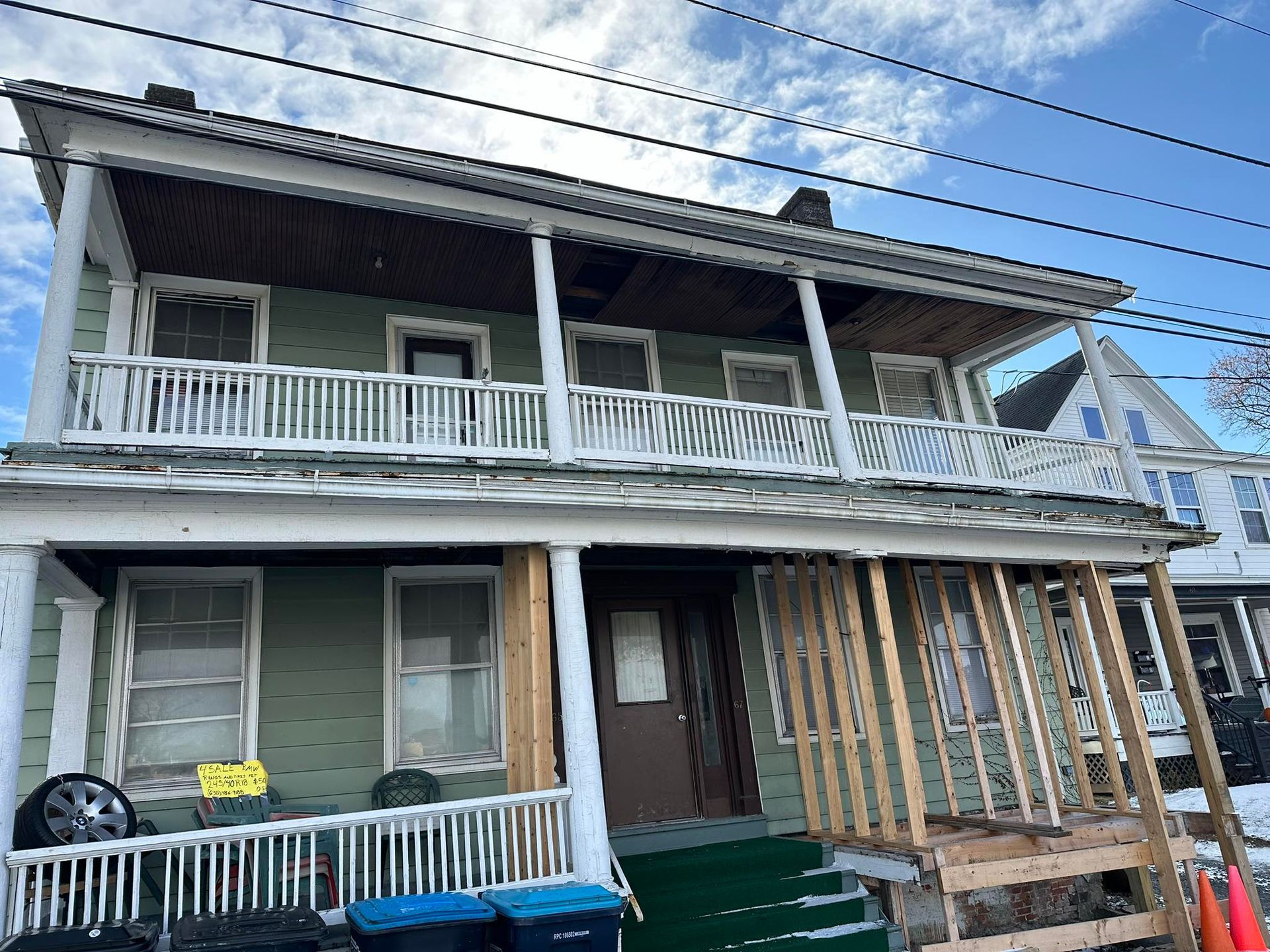 Two-story green house with white railings, brown door, and wooden porch undergoing repairs.