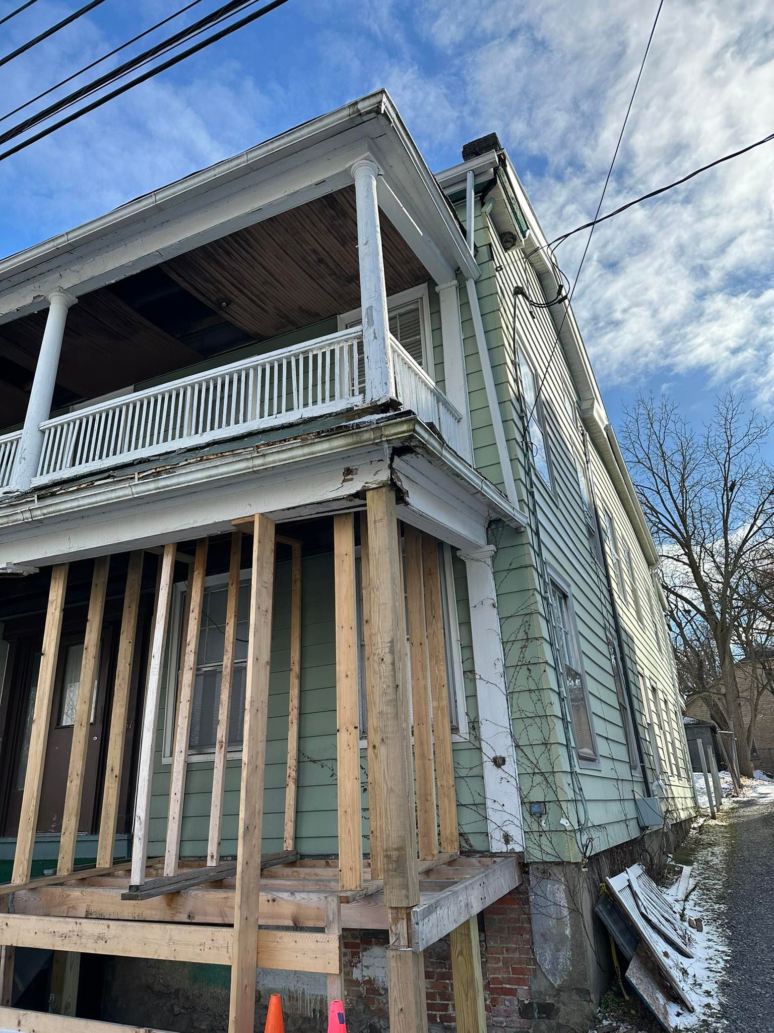 Two-story house with a porch under renovation; light green siding, weathered wood supports, and cloudy sky.