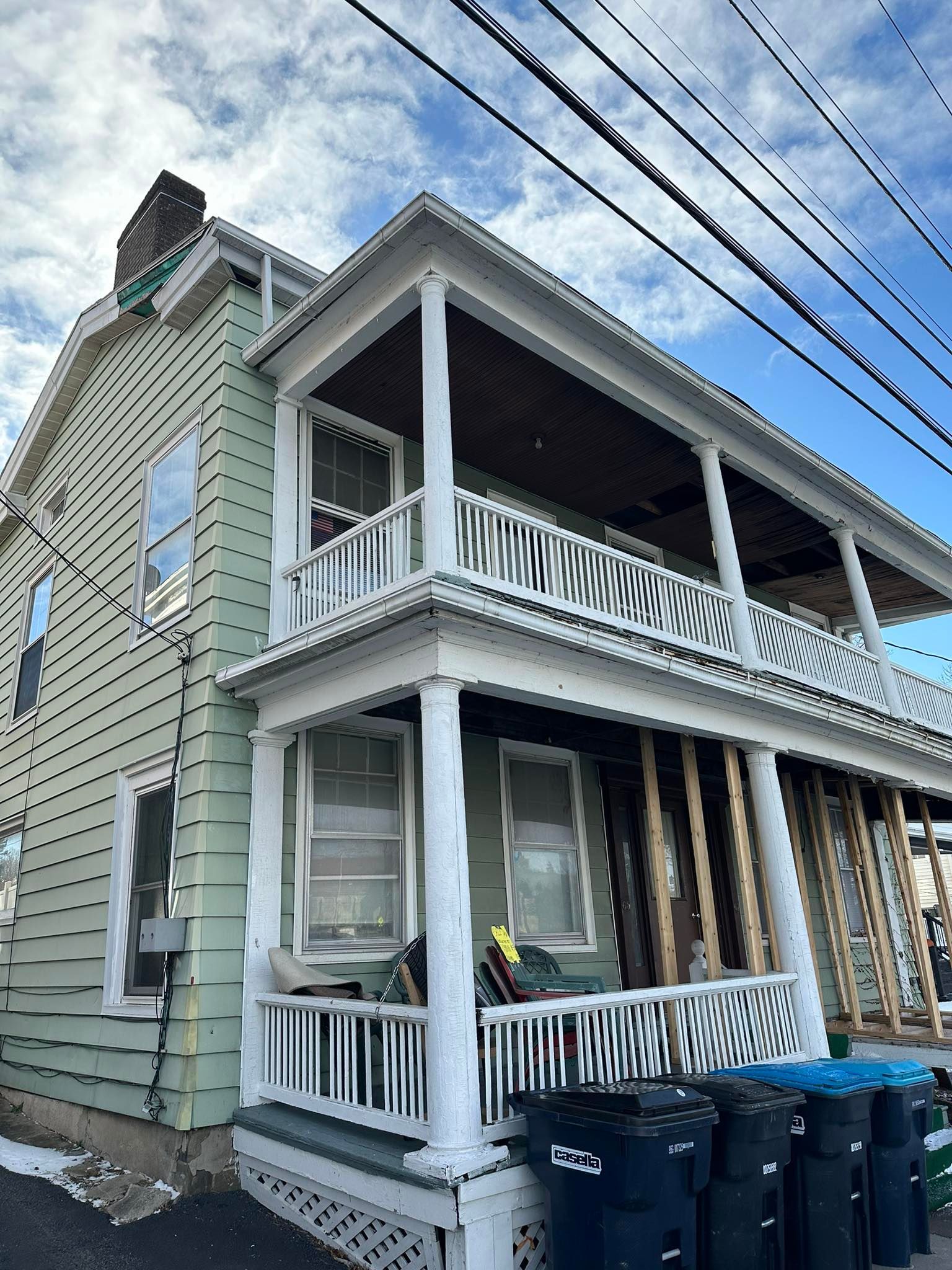 Two-story house with green siding and white trim, front porch under construction, blue sky with power lines.