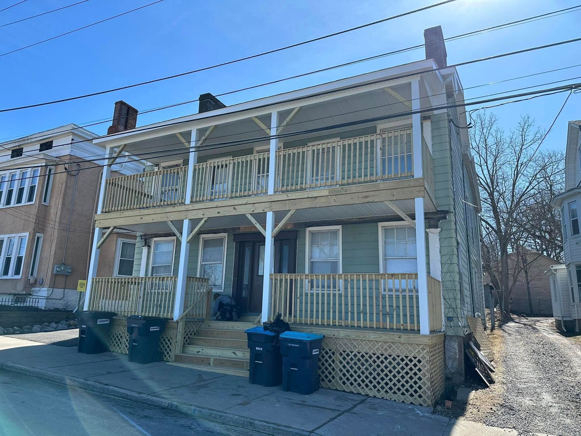 Two-story light green house with wooden porches, trash bins in front on a sunny day.