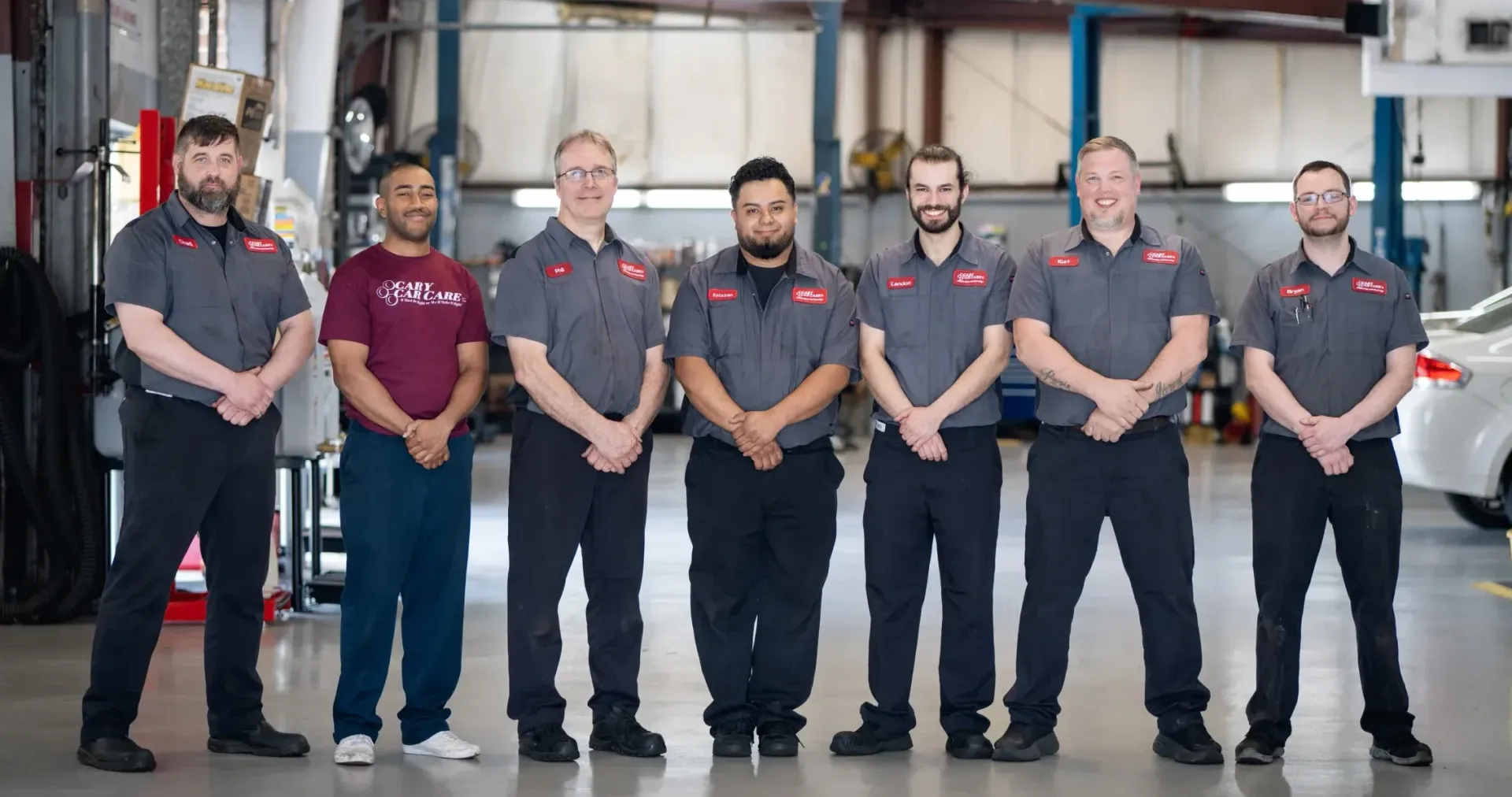 Seven auto mechanics in gray work shirts stand in a garage, arms crossed.