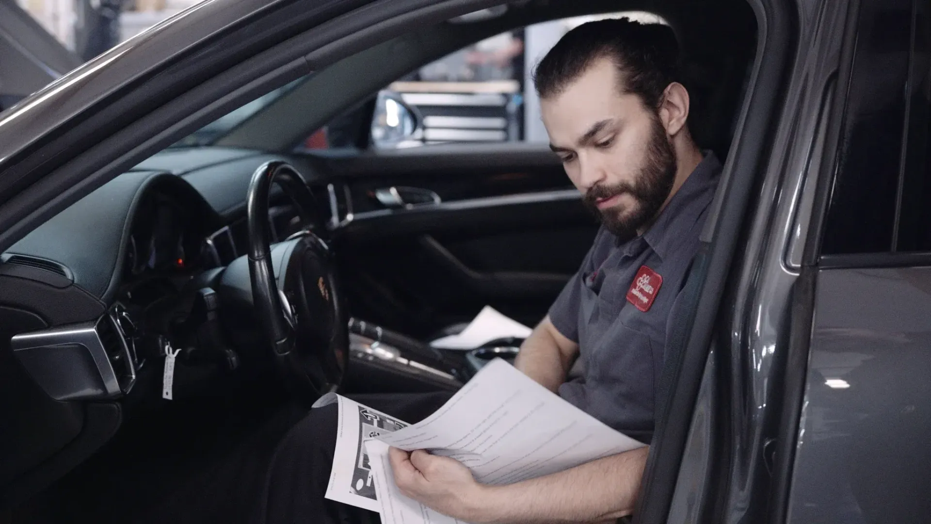 Mechanic inside car, reading paperwork. Shop setting; dark hair, beard, focused expression.