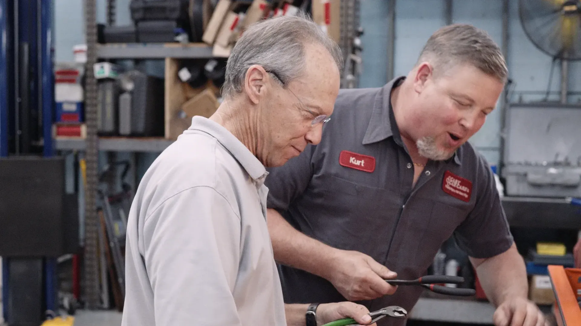 Man in a gray shirt looks at a tool held by a man in a red-and-gray uniform in a garage.