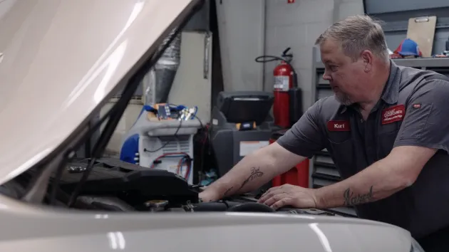 Mechanic working on car engine in a garage; gray shirt, open hood.