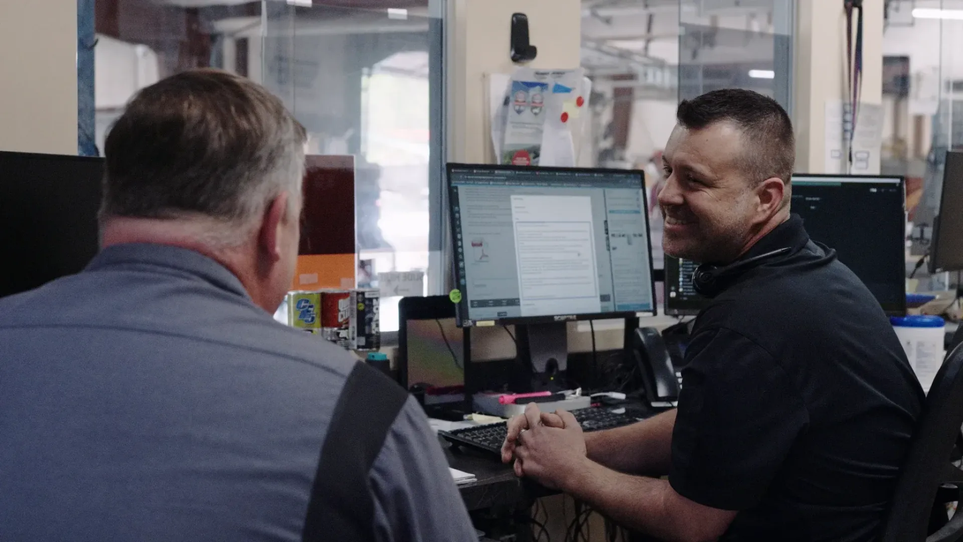 Two men at a desk; one facing away, the other smiling at the computer screen. Office setting with computer monitors.