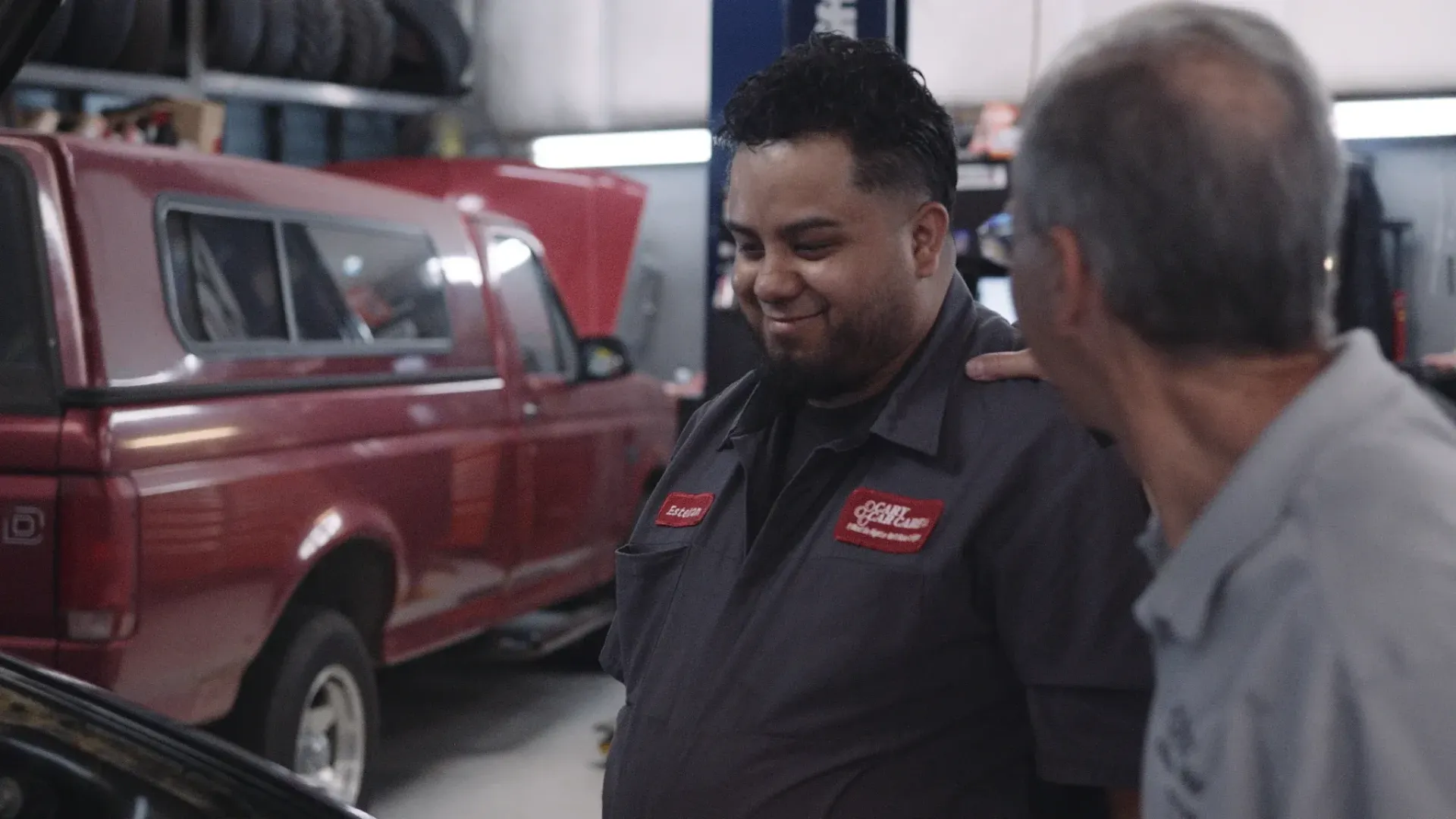 Man in gray mechanic uniform smiles, arm around another man's shoulder in auto shop. Red truck.