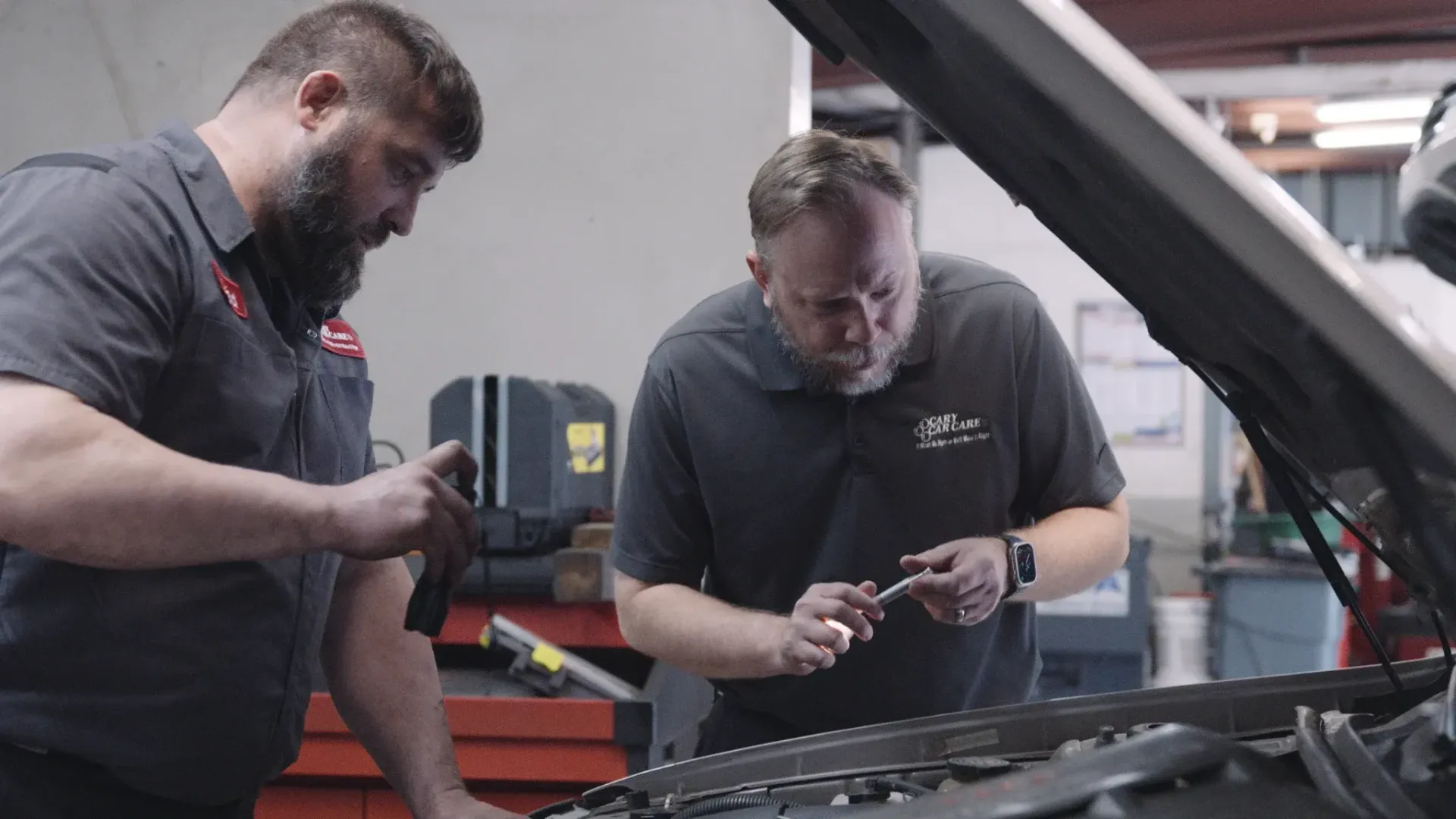 Two mechanics working on a car engine with tools in an auto repair shop.