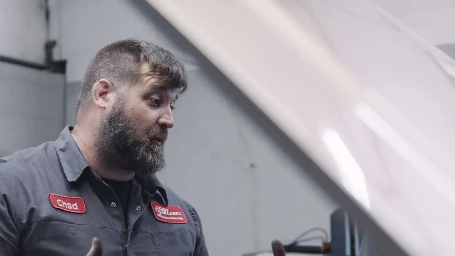 Mechanic in blue work shirt, gesturing toward car engine. Shop setting.