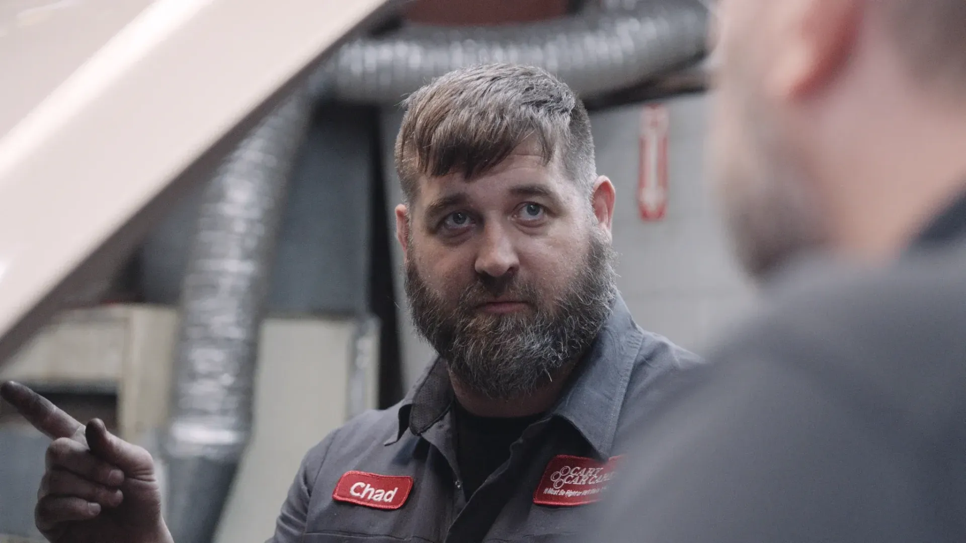 Mechanic pointing at car engine, talking to another mechanic in a garage setting.