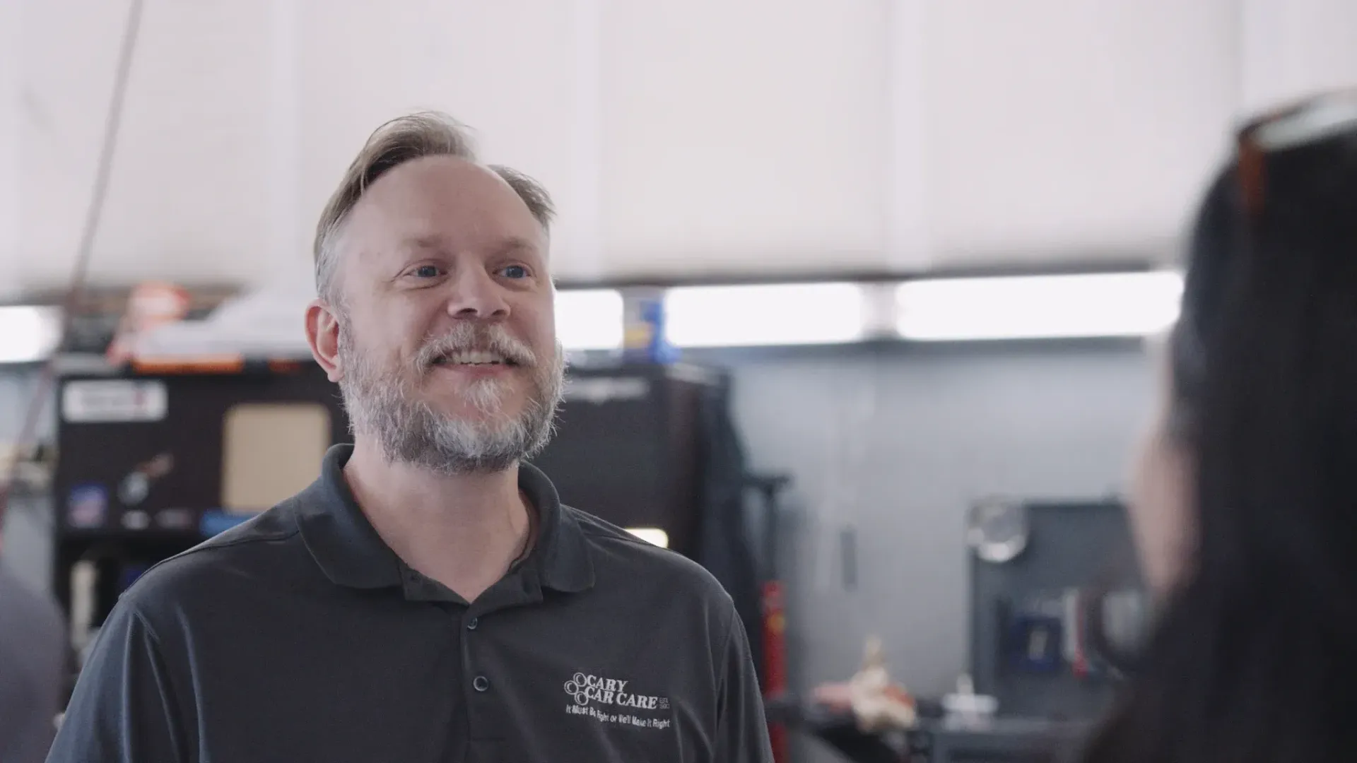 Man in a workshop smiles, talking to a person out of frame. He wears a dark polo shirt.