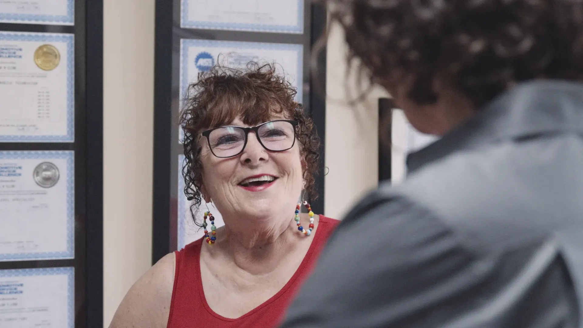Woman with glasses smiles at someone out of frame, in front of framed certificates.