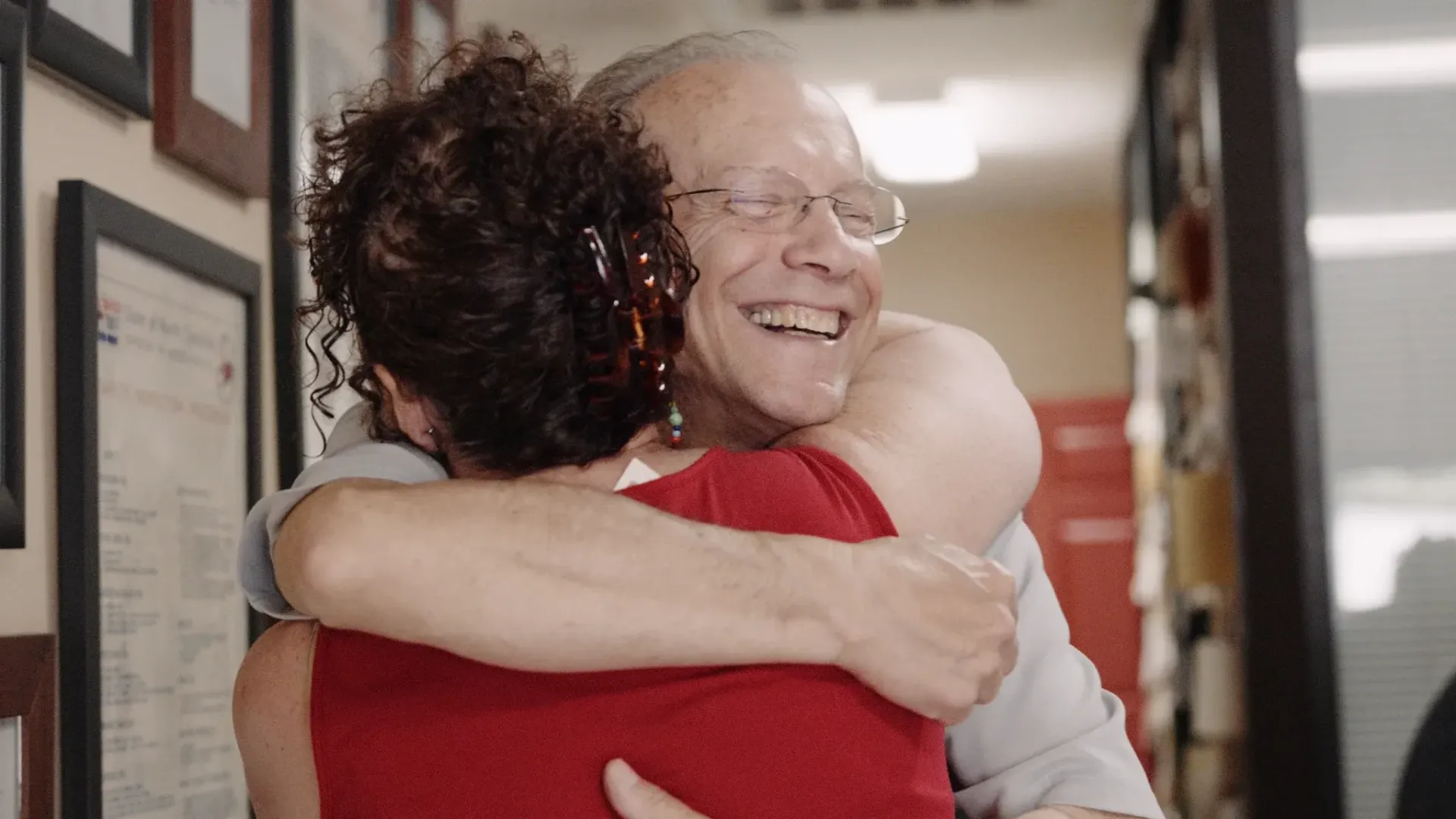 Man and woman embracing indoors; both smiling; wall with framed documents.