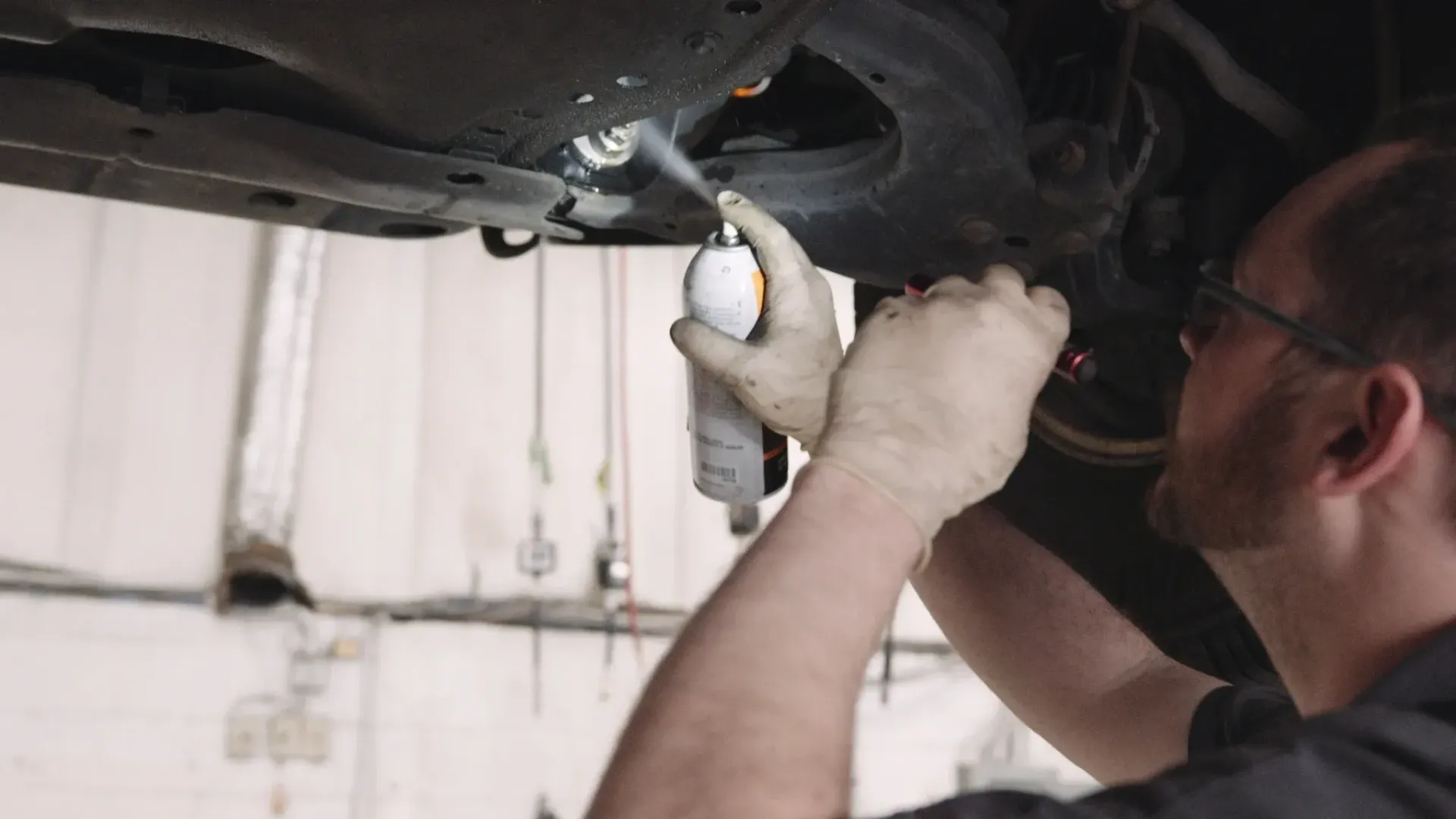 Mechanic sprays undercarriage of a car. Wearing gloves and glasses, working in a shop.