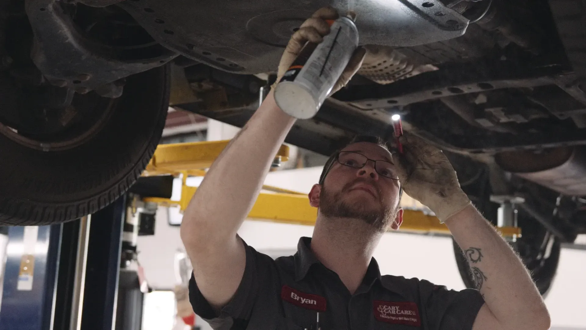 Mechanic sprays fluid under a car, using a flashlight.