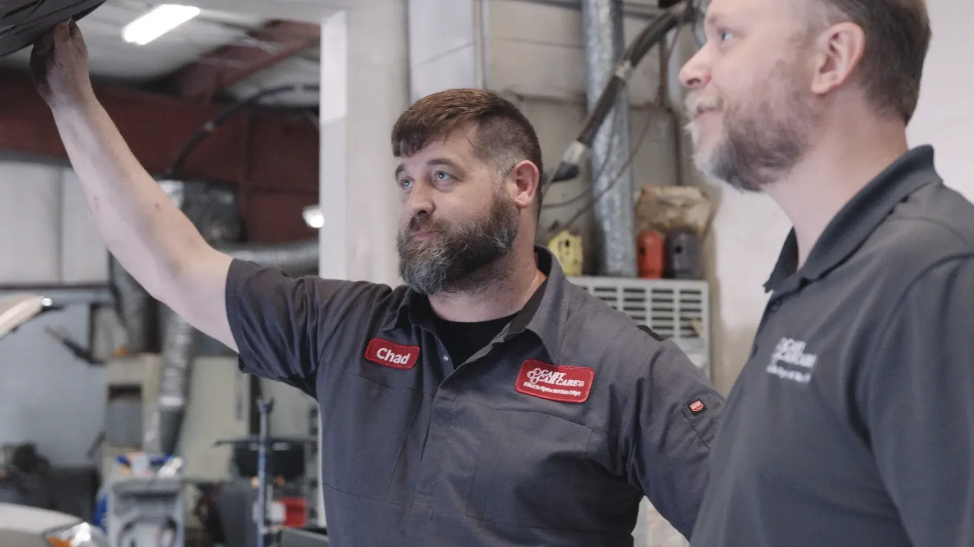 Two men in a repair shop examine the underside of a vehicle. One points, the other looks on, both wearing work shirts.