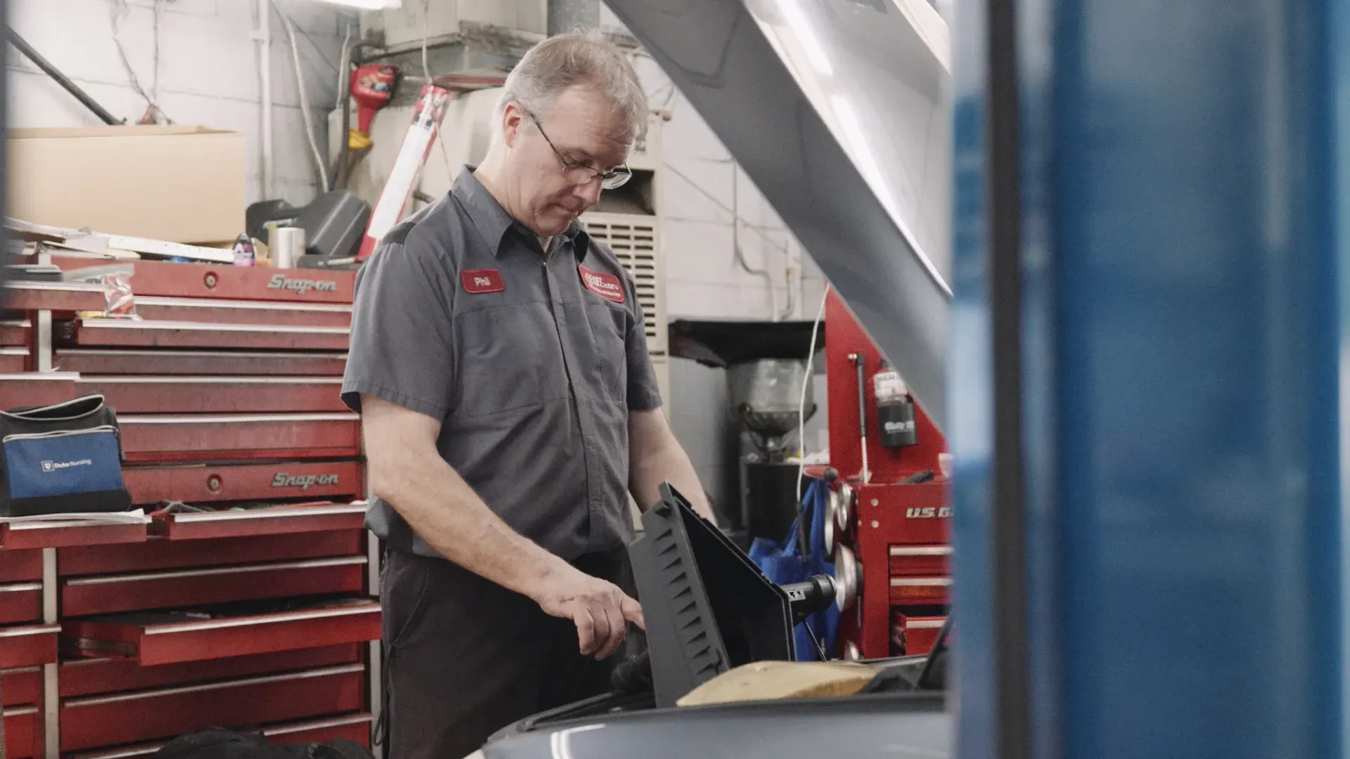 Mechanic working on a car in a shop, looking under the hood. He wears a gray shirt and glasses. Red toolbox in background.