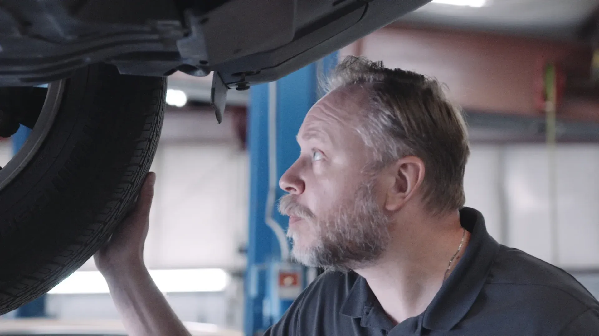 Mechanic examines a car tire from below in a garage, looking intently.