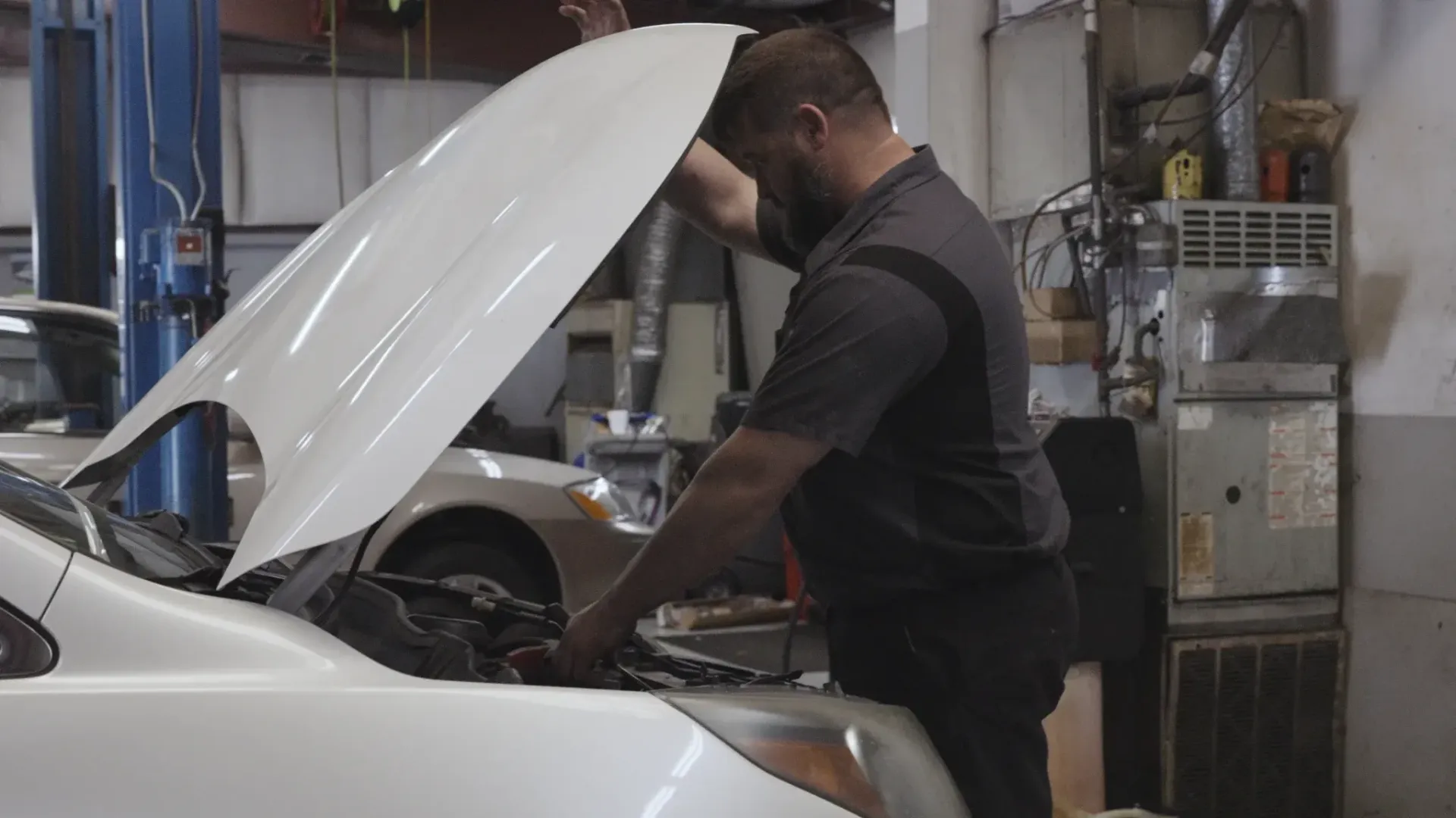Mechanic in a gray shirt inspecting a white car with hood open in a garage.