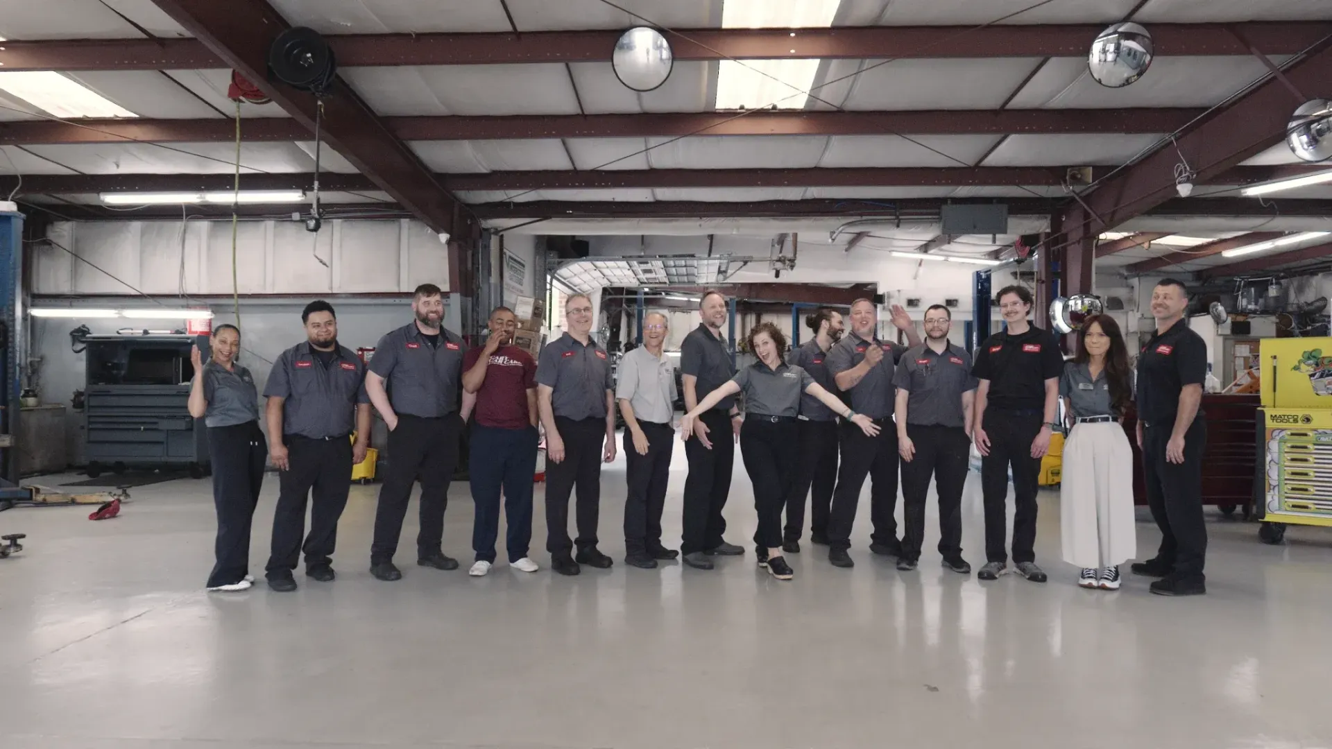 Group of people in an auto repair shop; posing with arms out. Various uniforms, setting is a garage with high ceilings.