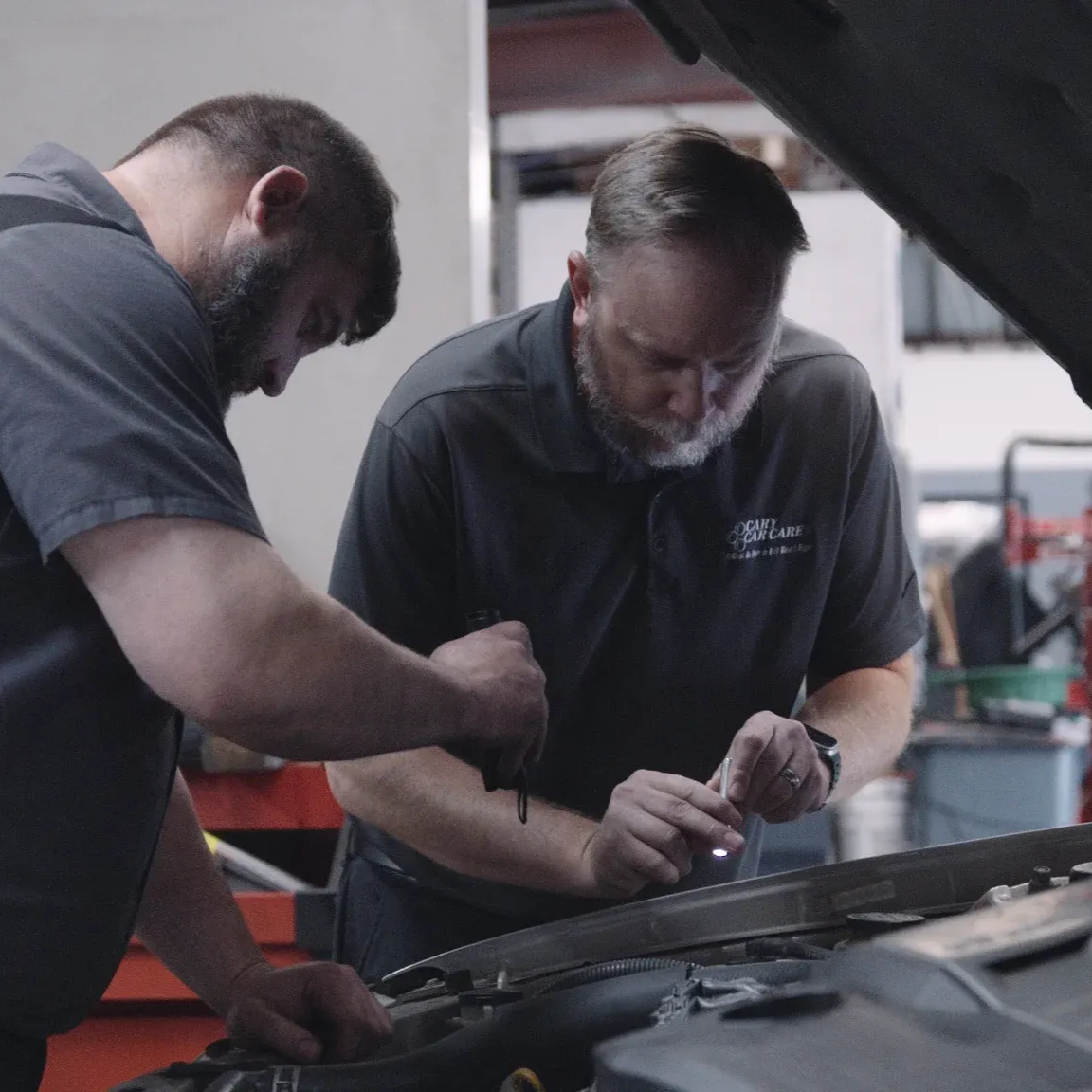 Two mechanics examining a car engine in a shop. One holds a flashlight, the other works.