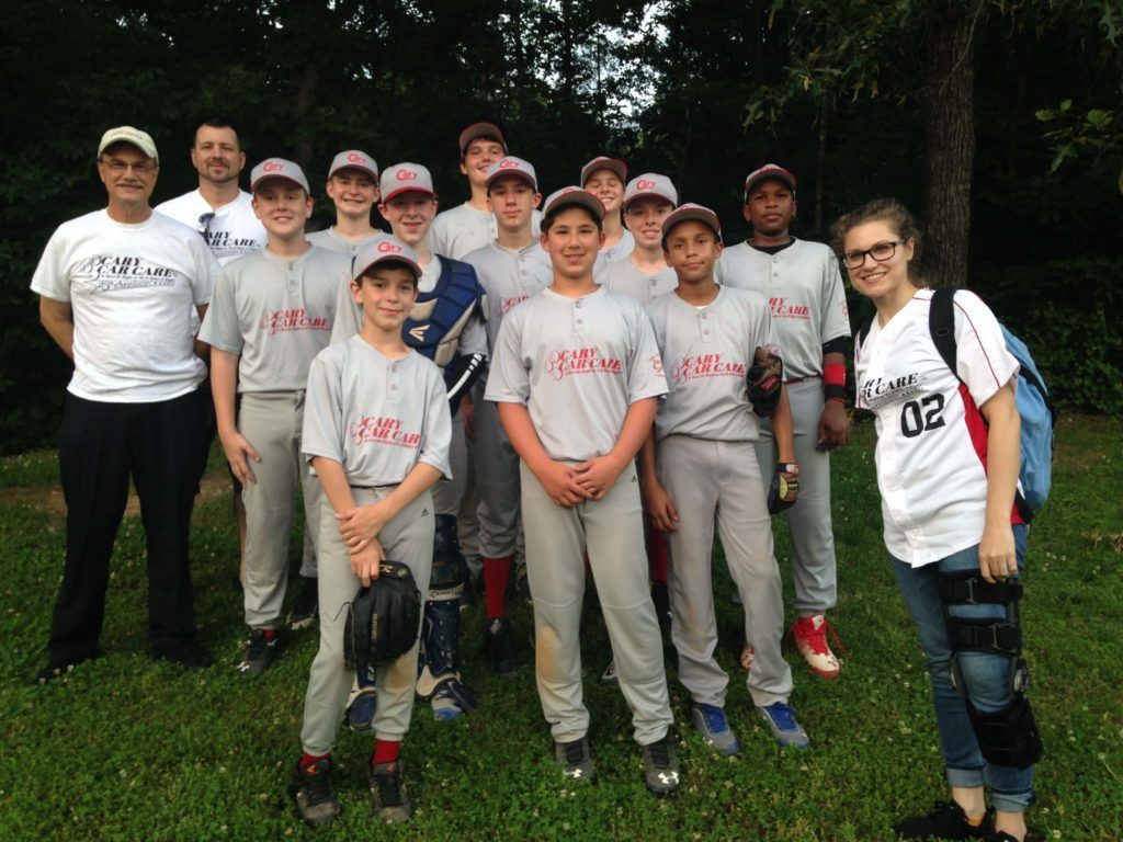 Baseball team in gray uniforms, posed outdoors on grass; two coaches, teen players, and a person with a leg brace.