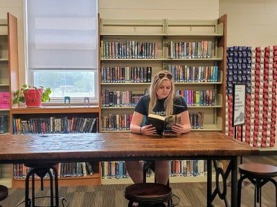 Woman reading a book at a table in a library with bookshelves and an American flag display.