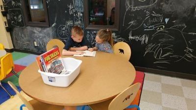 Two children reading at a round table in a classroom with a chalkboard wall.