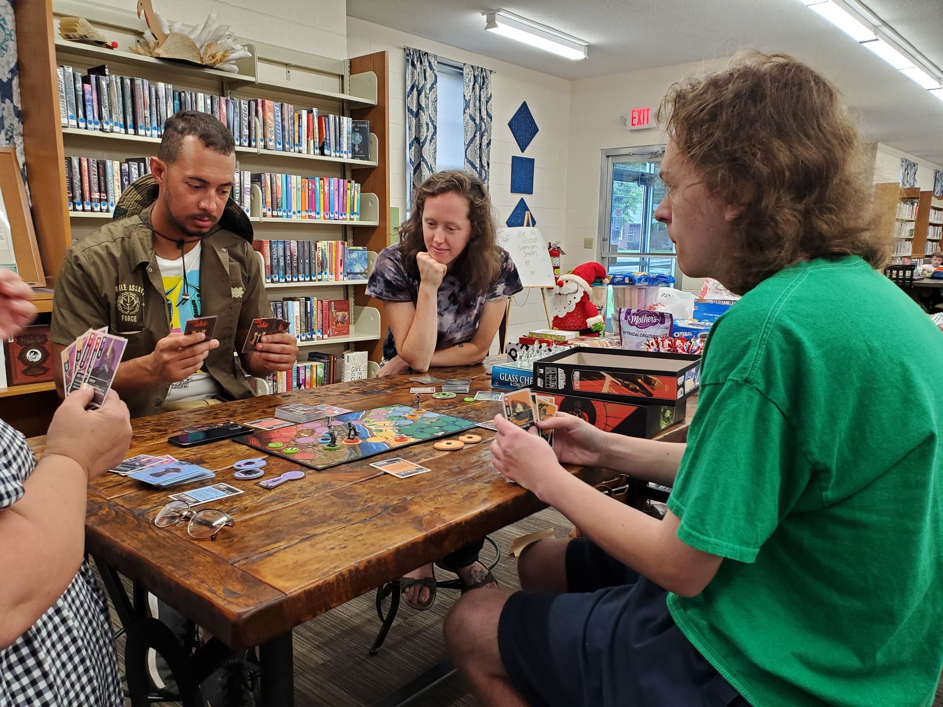 People playing a board game at a table in a library. Cards and game pieces are visible.