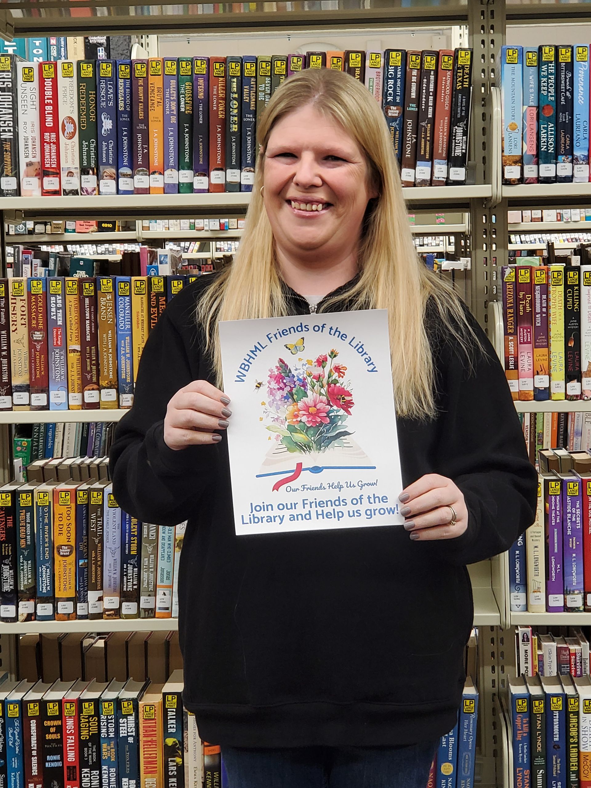 Woman in a library holding a drawing of flowers. She is smiling. Behind her are shelves of VHS tapes.
