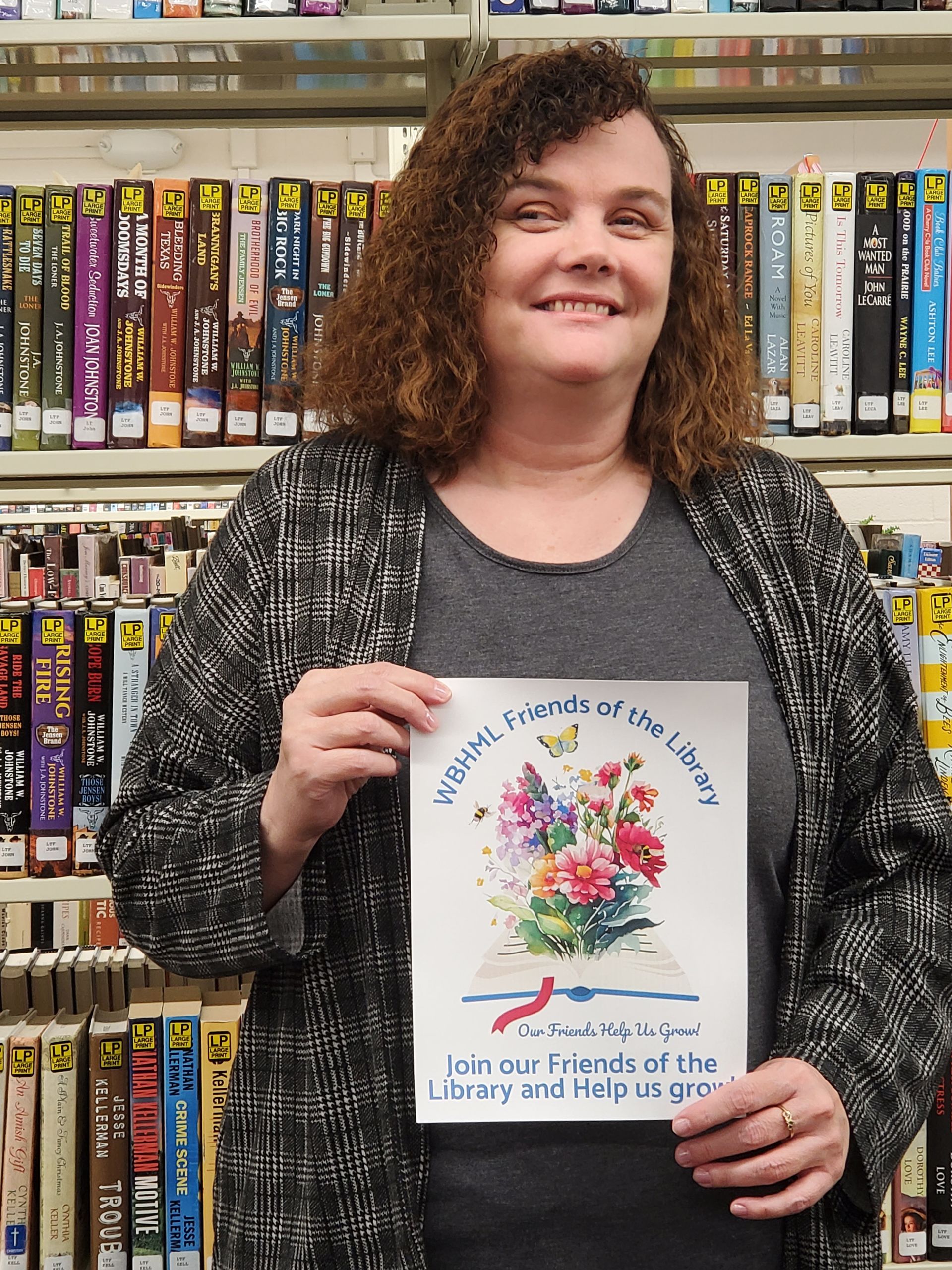 Woman in a library holding a sign with flowers and text; smiling. Shelves with videos are in the background.