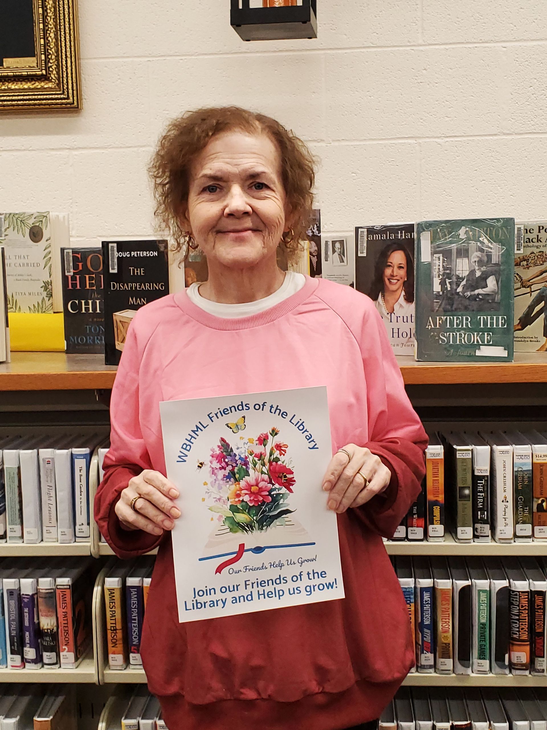 Woman in pink sweater holds a colorful floral sign in a library.