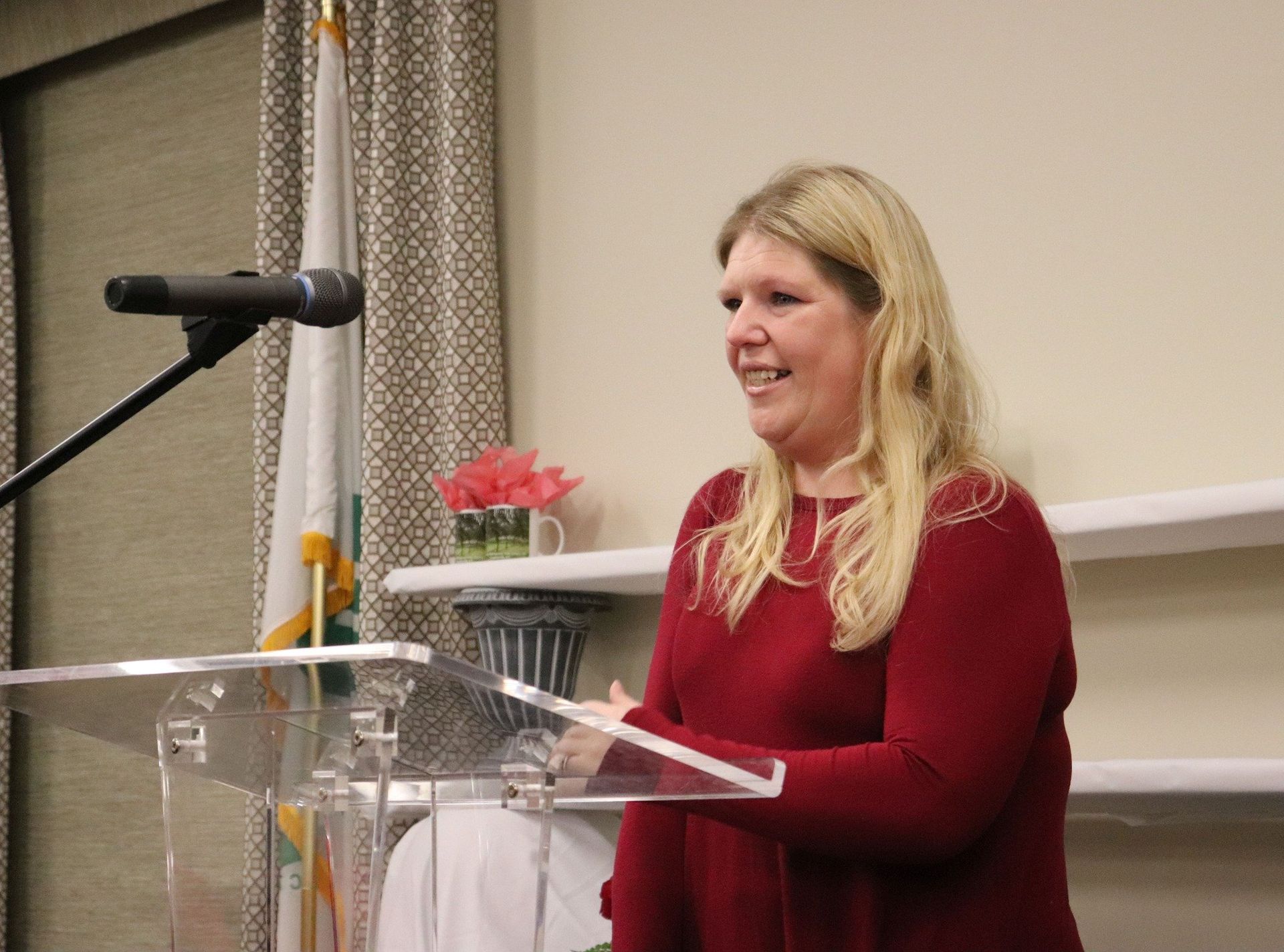 Woman speaking at a podium, smiling. Wearing a red sweater. Microphone and flags in the background.