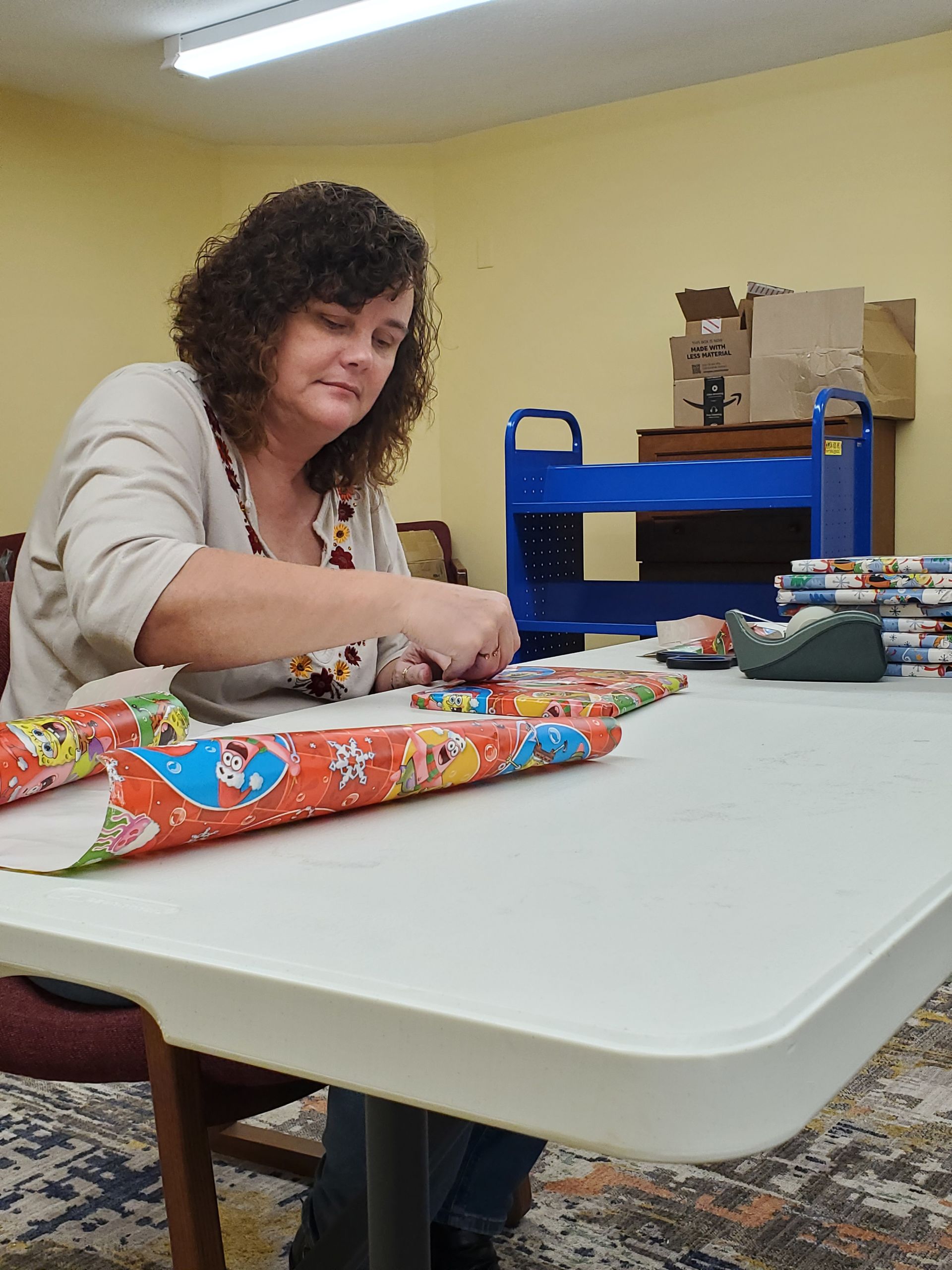 Woman wrapping presents at a table; colorful wrapping paper, tape dispenser, blue cart, boxes in the background.