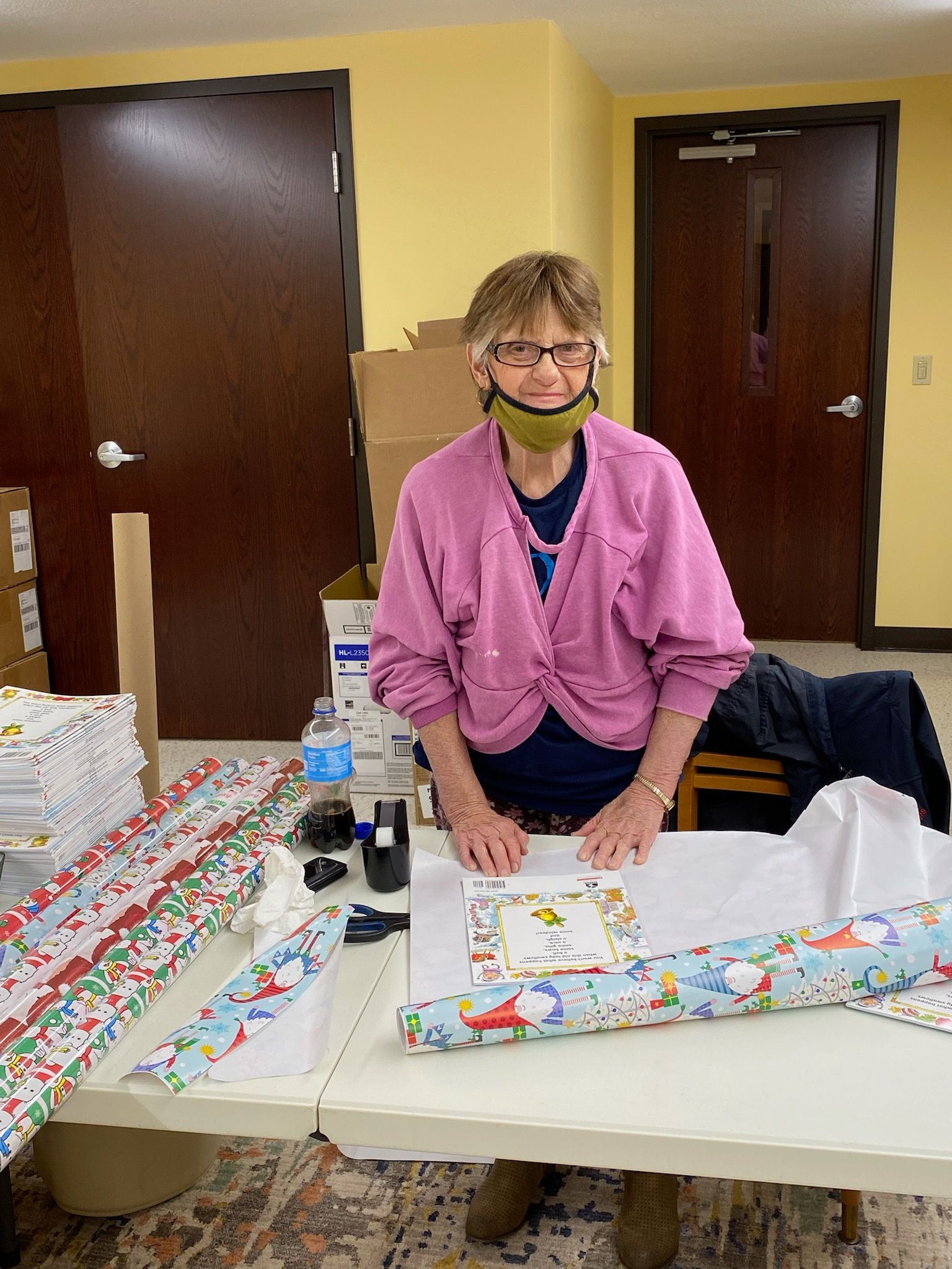 Woman wearing a face mask wrapping gifts at a table. Wrapping paper and supplies are visible.