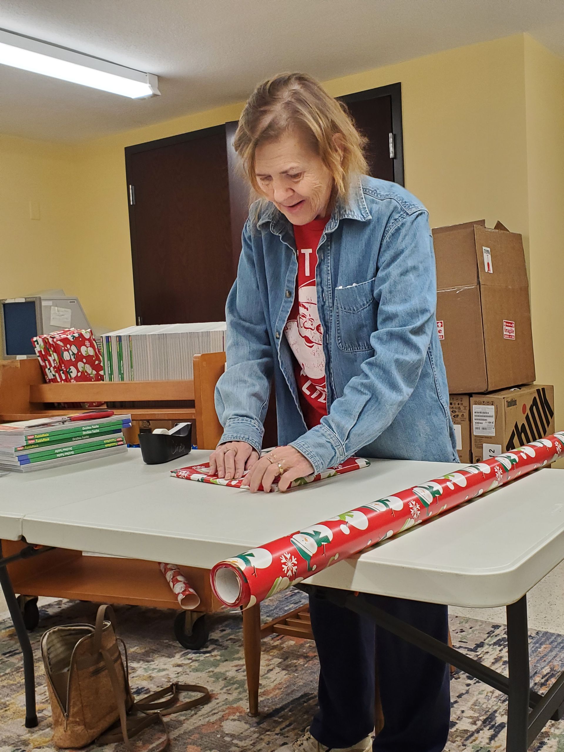 Woman wrapping a gift on a table; red wrapping paper, casual clothing, indoor setting.