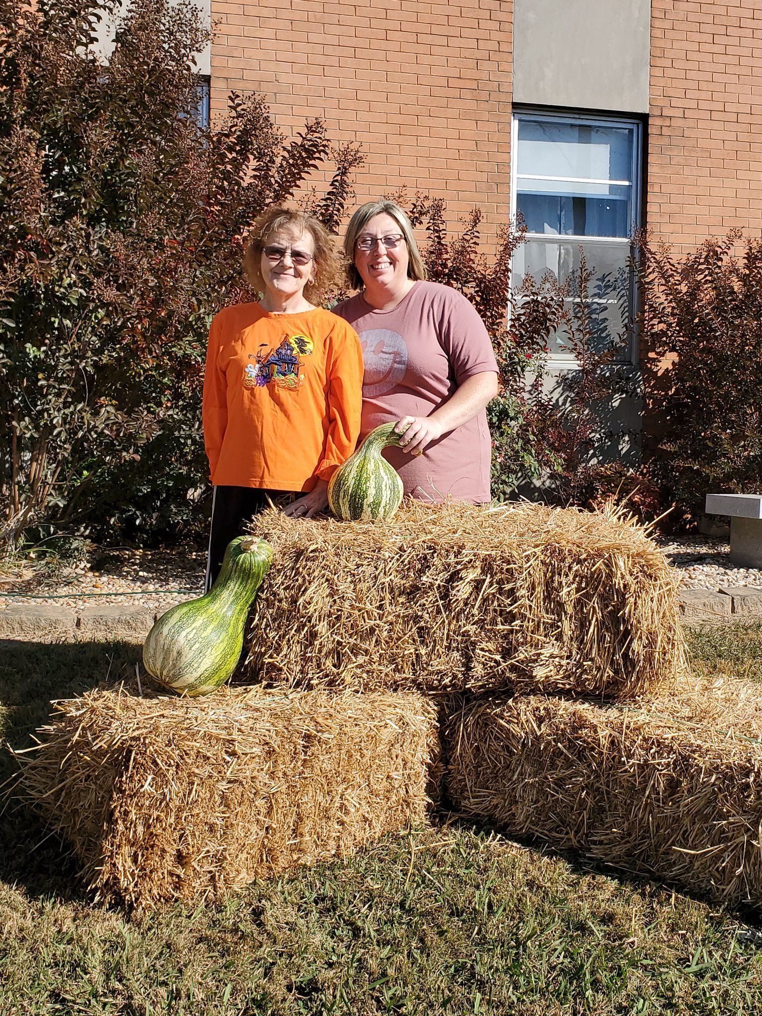 Two women pose behind hay bales with decorative gourds outside a brick building.
