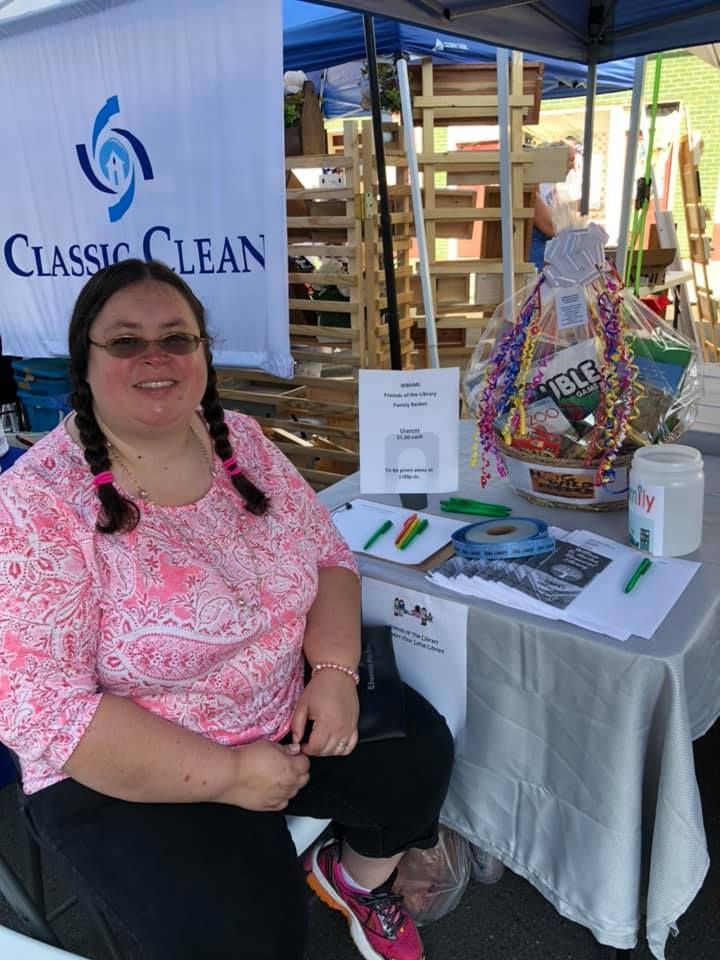 Woman at a Classic Clean booth, smiling. Pink patterned shirt, braids, seated. Table with basket, brochures.