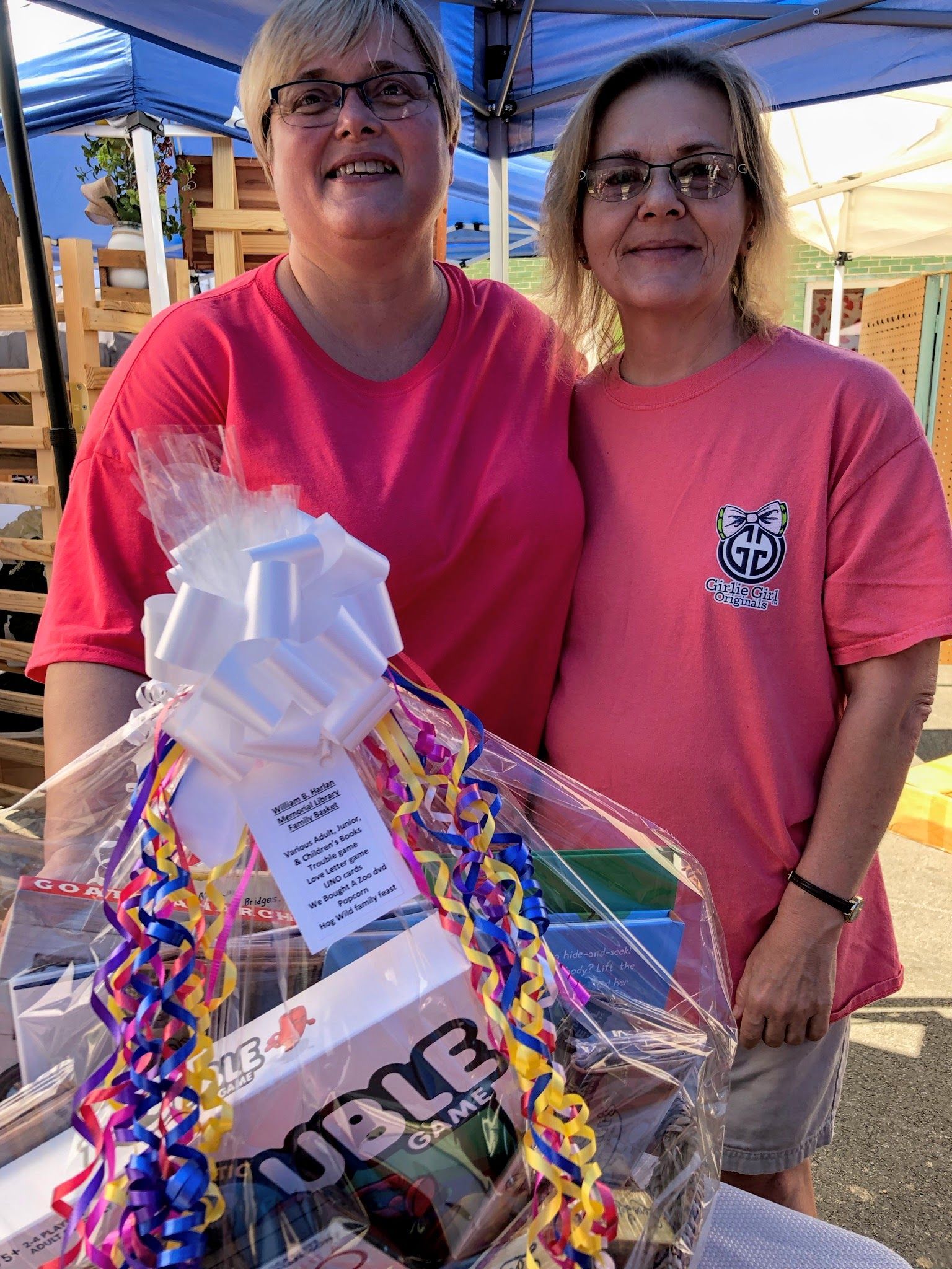 Two women stand behind a gift basket at an outdoor event, smiling.
