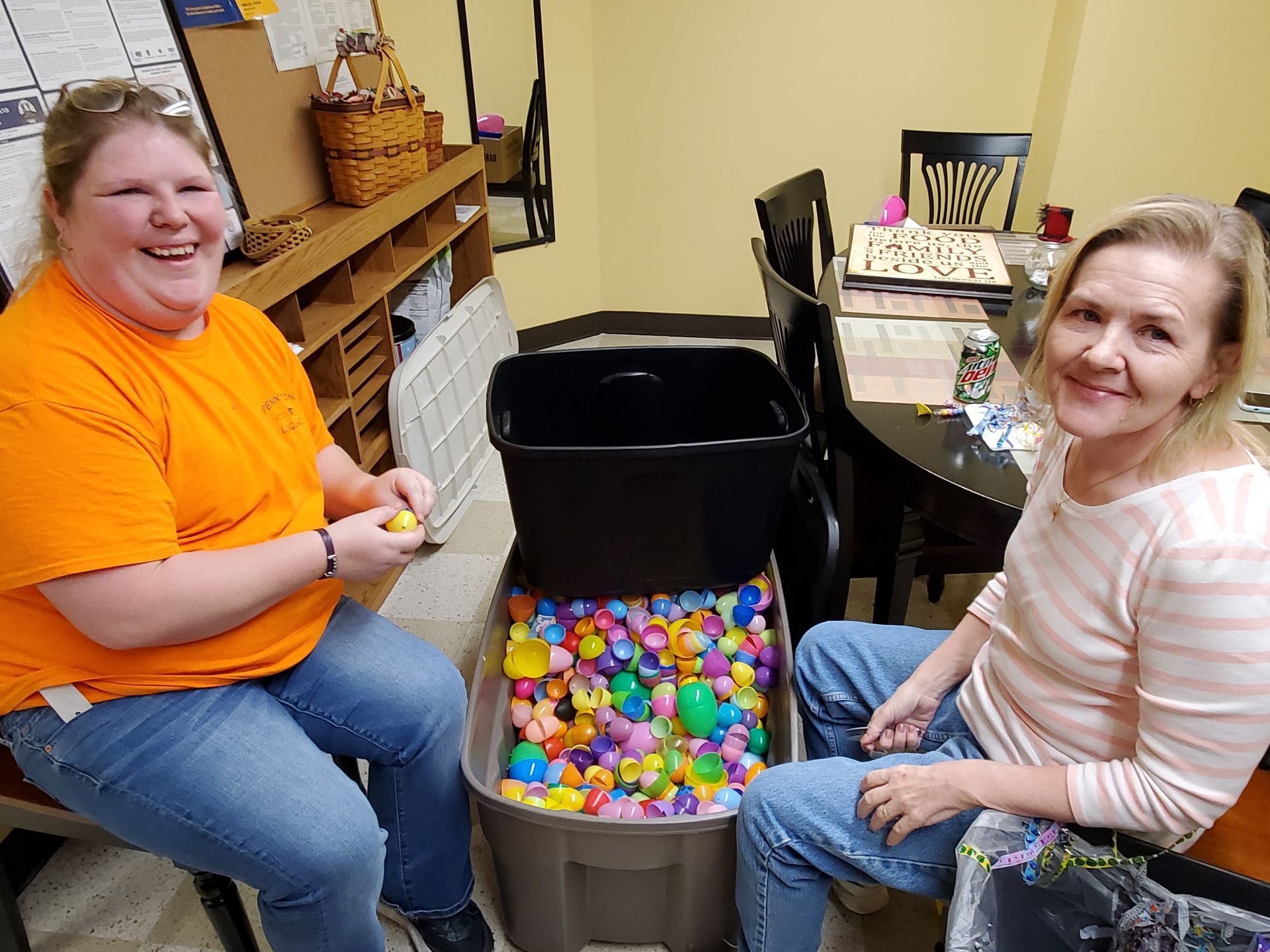 Two women preparing Easter eggs indoors; one in orange shirt laughs, other smiles. Eggs and basket visible.