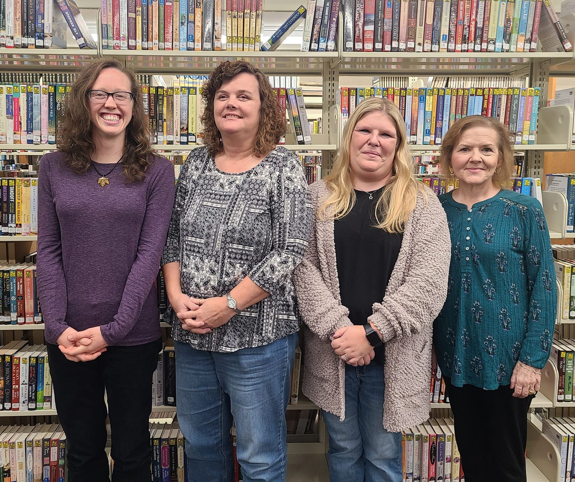 Four women standing in front of a bookshelf, smiling. One wears purple, the others wear patterned shirts and sweaters.